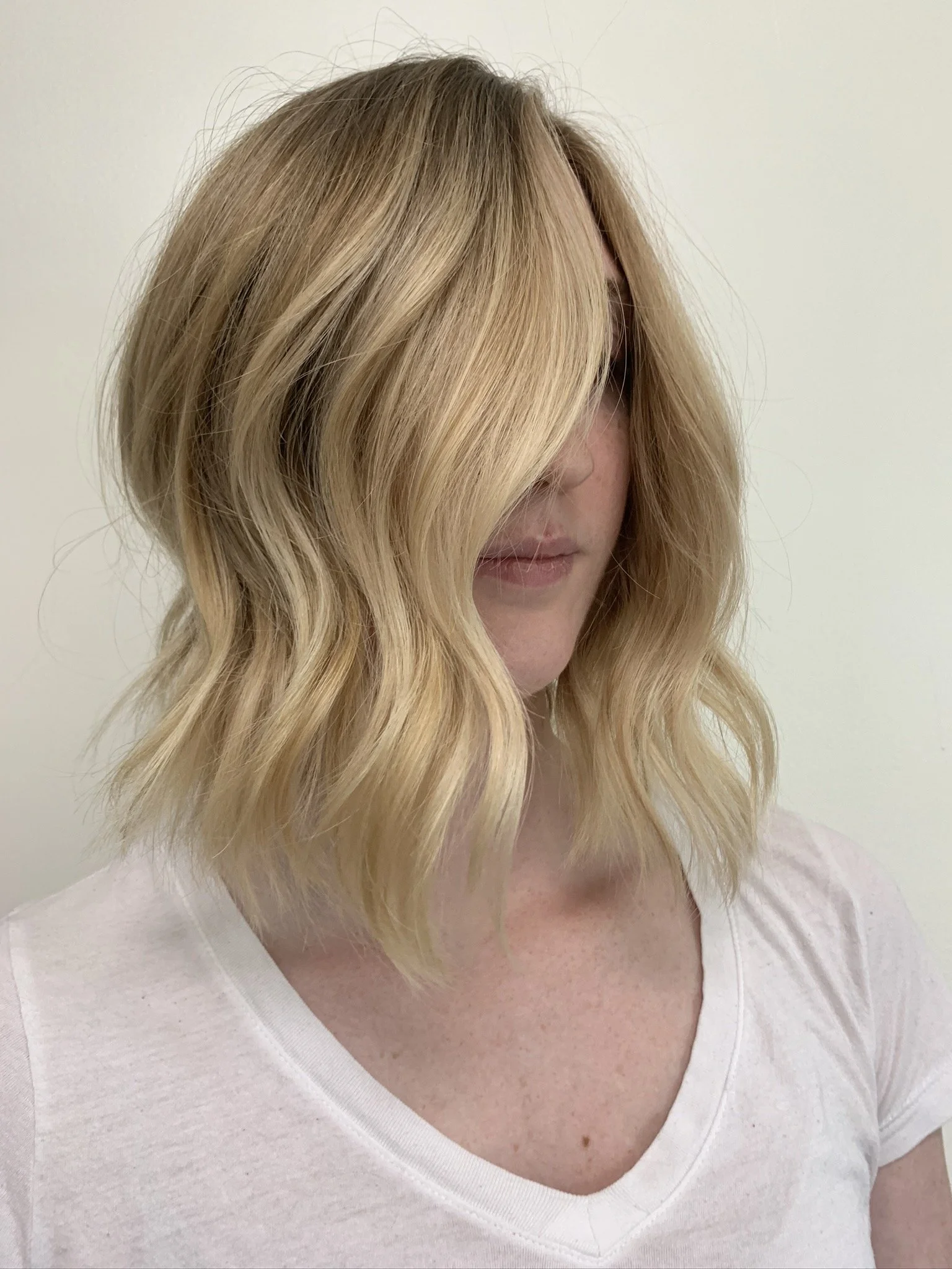 Woman with shoulder-length wavy blonde hair covering part of her face, wearing a white V-neck shirt, standing against a plain light-colored wall.