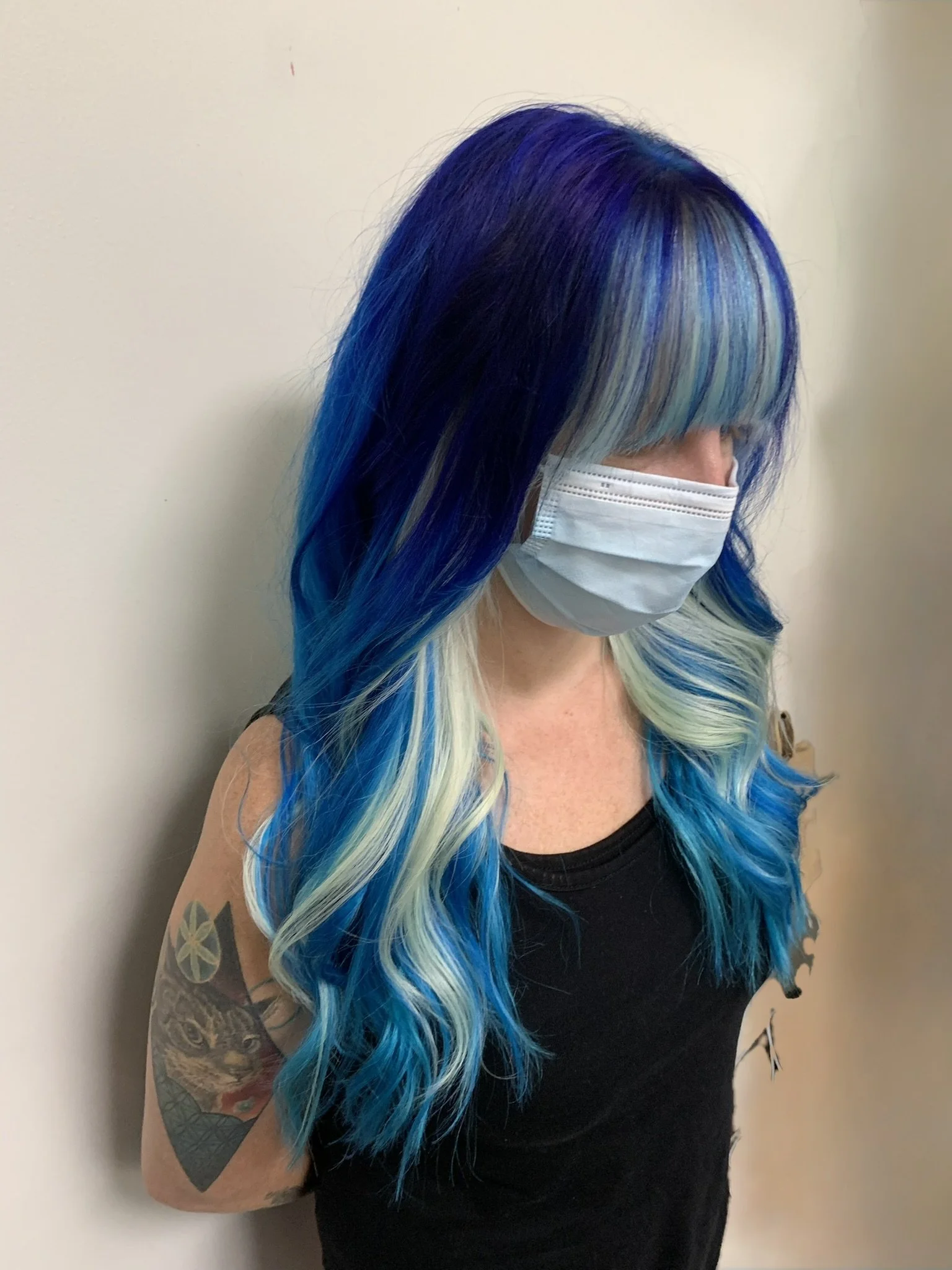 Woman with long, vibrant blue and light blue hair, wearing a gray face mask and black sleeveless top, standing against a beige wall.