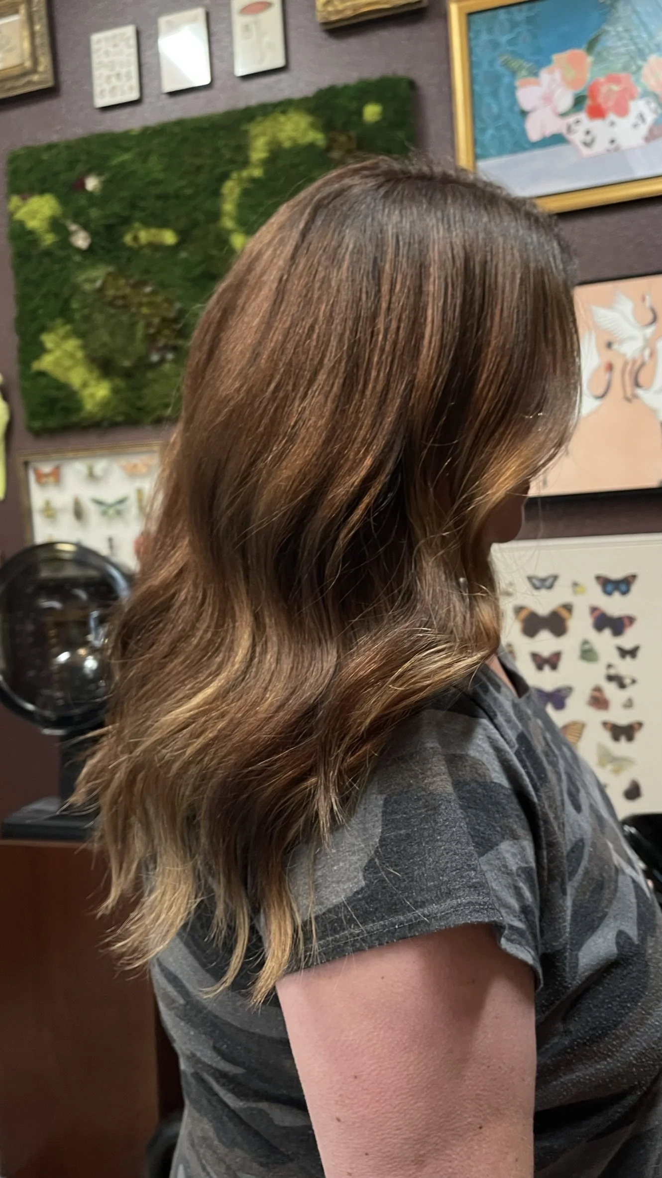 Side view of a woman with long, wavy brown hair in a salon, with butterfly and nature-themed artwork on the wall behind her.
