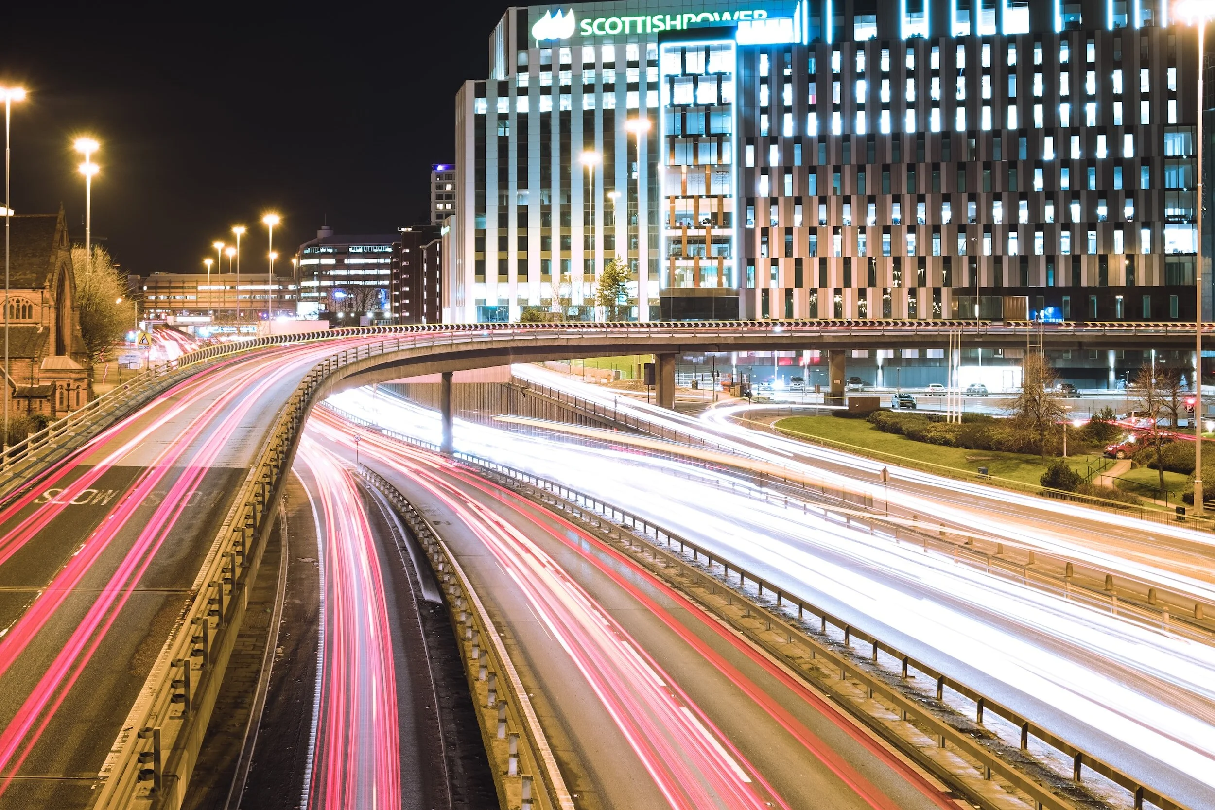 Nighttime cityscape with illuminated buildings and blurred car lights on the highway showing streaks of red and white light trails.