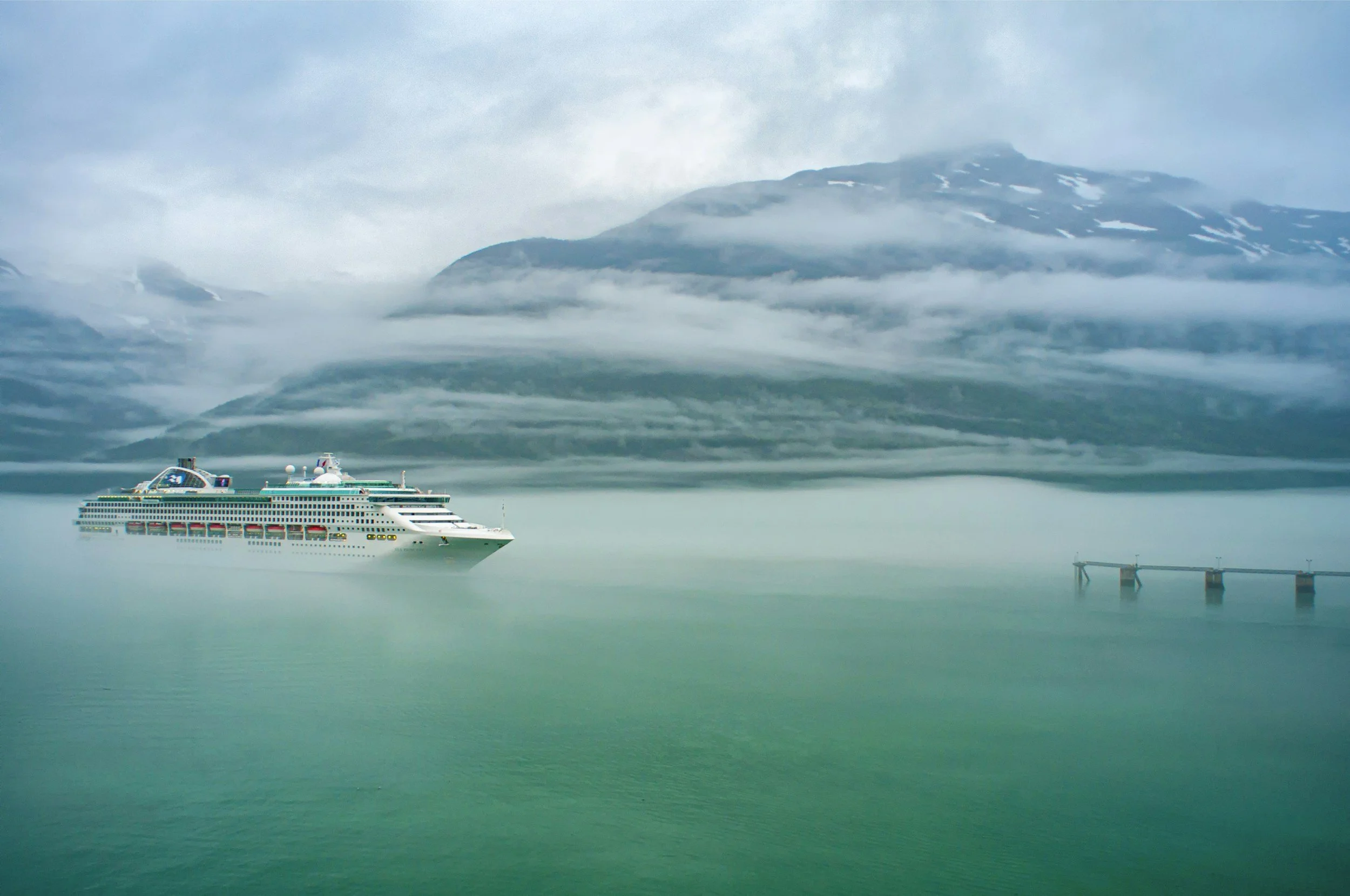 Cruise ship sailing in calm, greenish water with fog and mountains in the background.