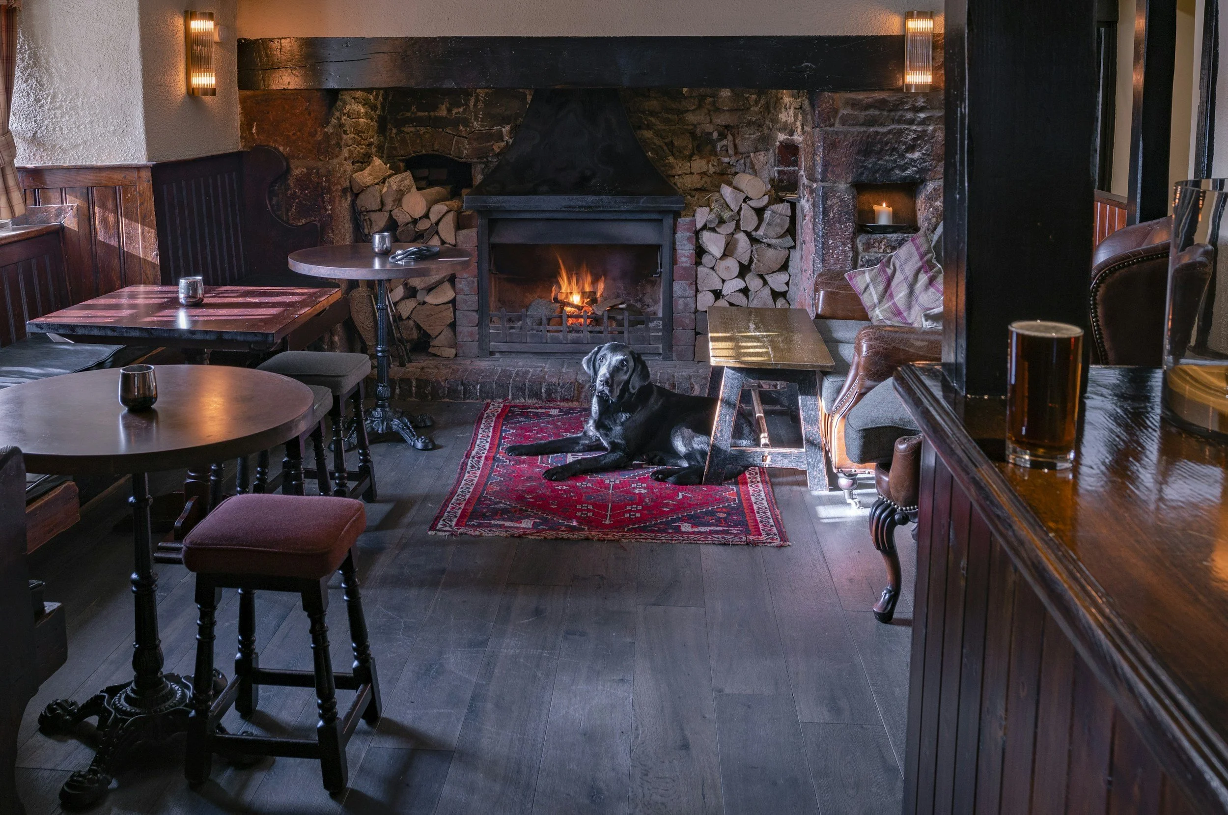 A cozy pub interior with a lit brick fireplace, stacked firewood on either side, and a black dog lying on a red patterned rug in front of the fireplace. The room has wooden tables, some with small candles, and comfy chairs and booths.