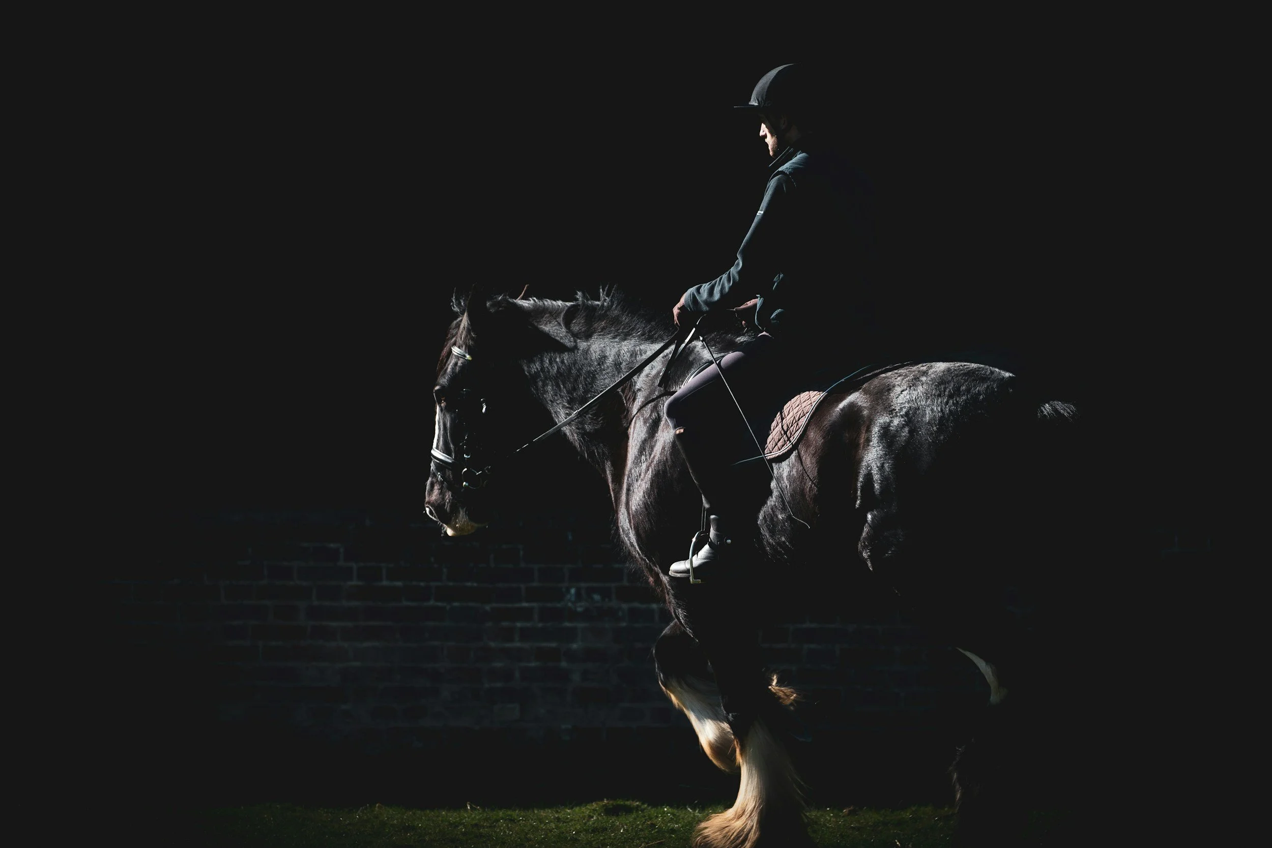 A person riding a horse in a dark setting, with a black brick wall in the background. The rider is wearing a helmet and equestrian attire.