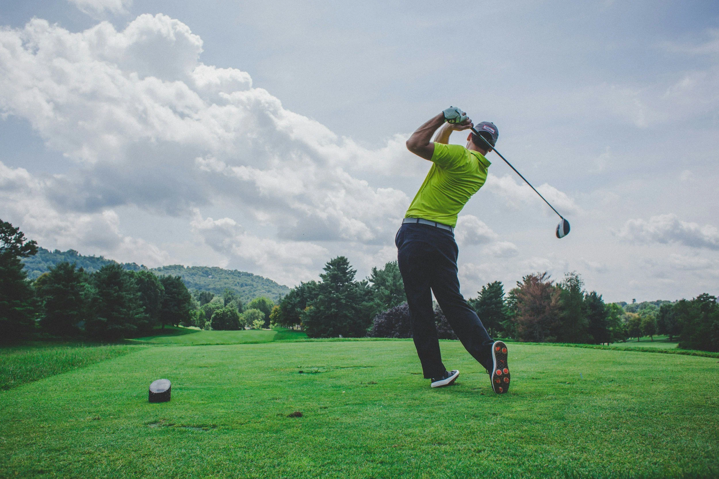 A man in a bright green shirt and dark pants hitting a golf ball on a golf course, with a scenic landscape and cloudy sky in the background.