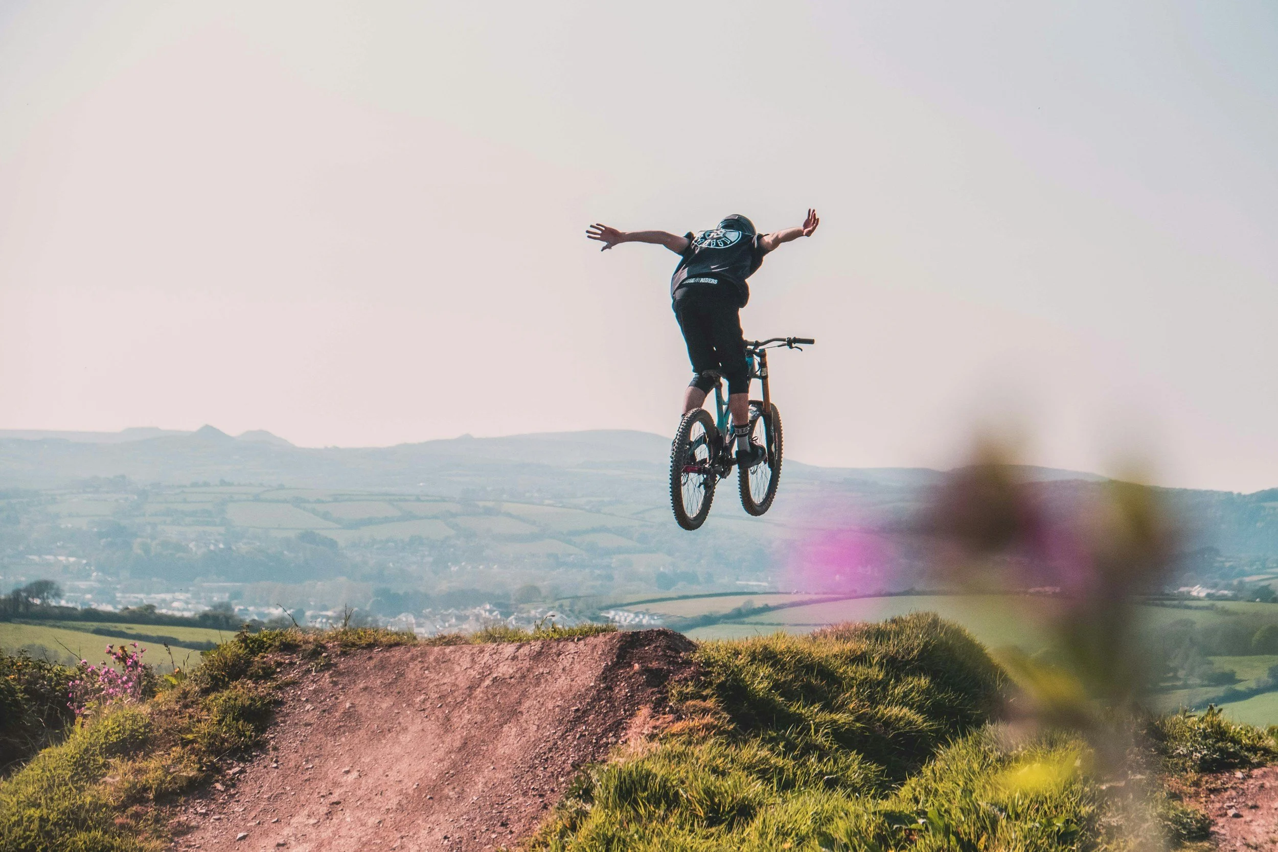 A person performing a jump on a mountain bike off a dirt ramp in a grassy landscape with rolling hills in the background.