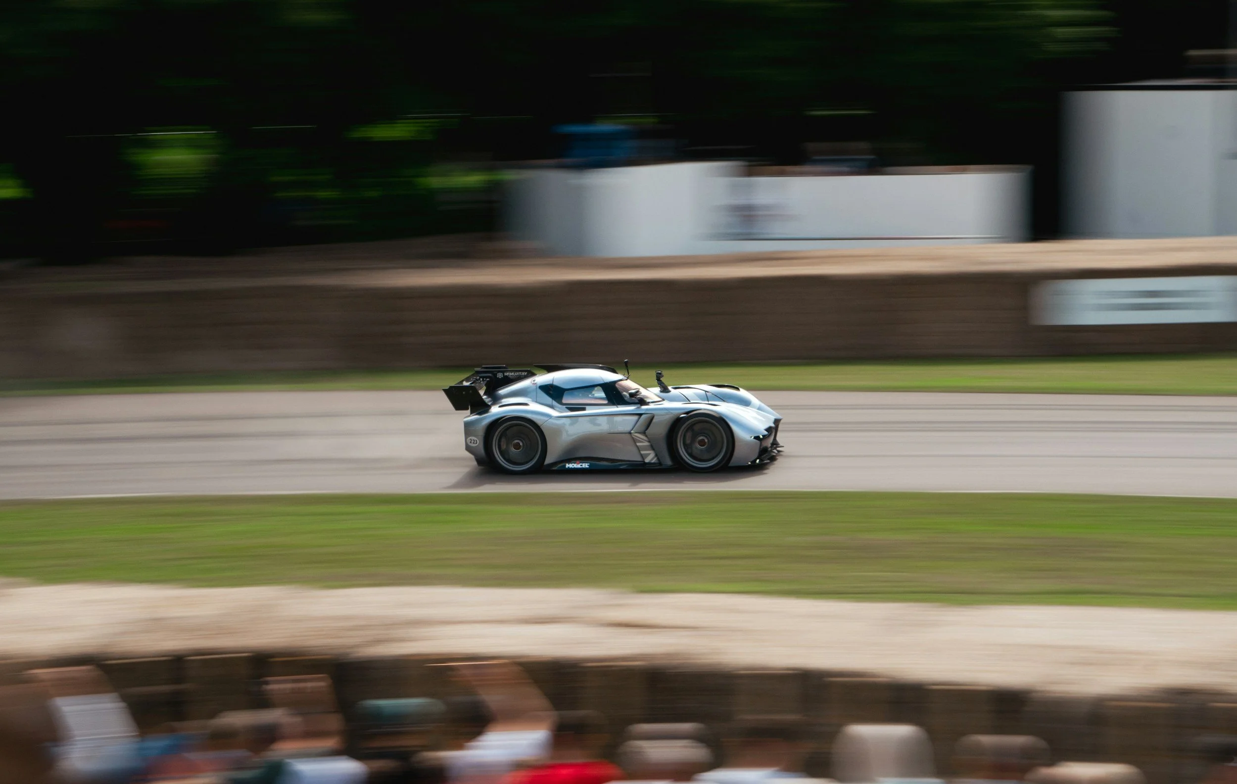 A sleek, silver race car speeding on a racetrack with a blurred background and spectators in the foreground.