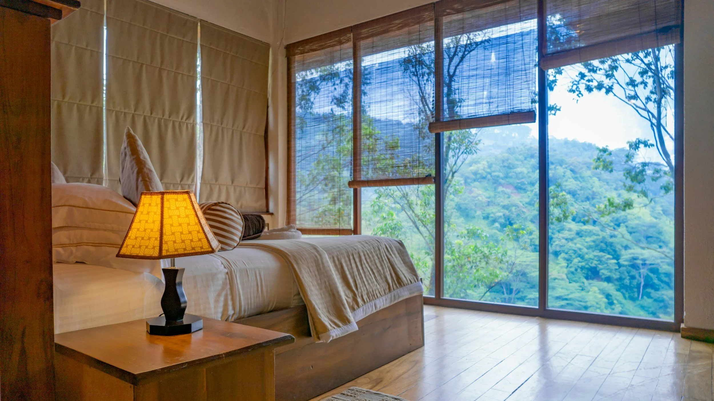 A cozy bedroom with a wooden bed frame, white bedding, and pillows, illuminated by a bedside lamp. Large windows with bamboo blinds reveal a lush green forest outside.
