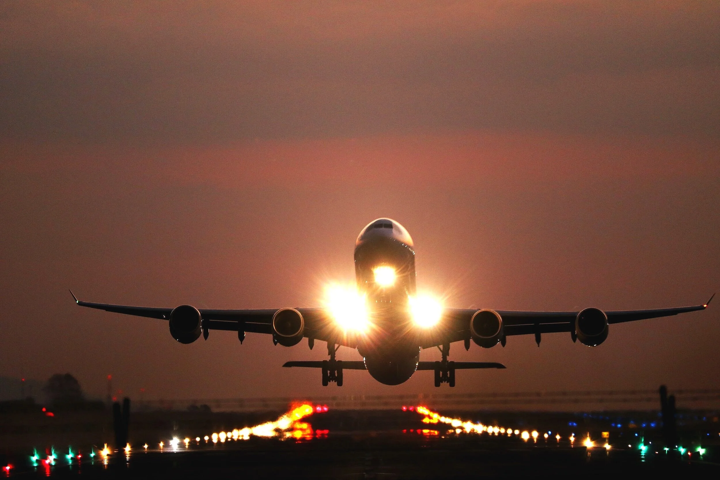 A commercial airplane during sunset or sunrise, with its headlights on, taking off from a runway illuminated with runway lights.
