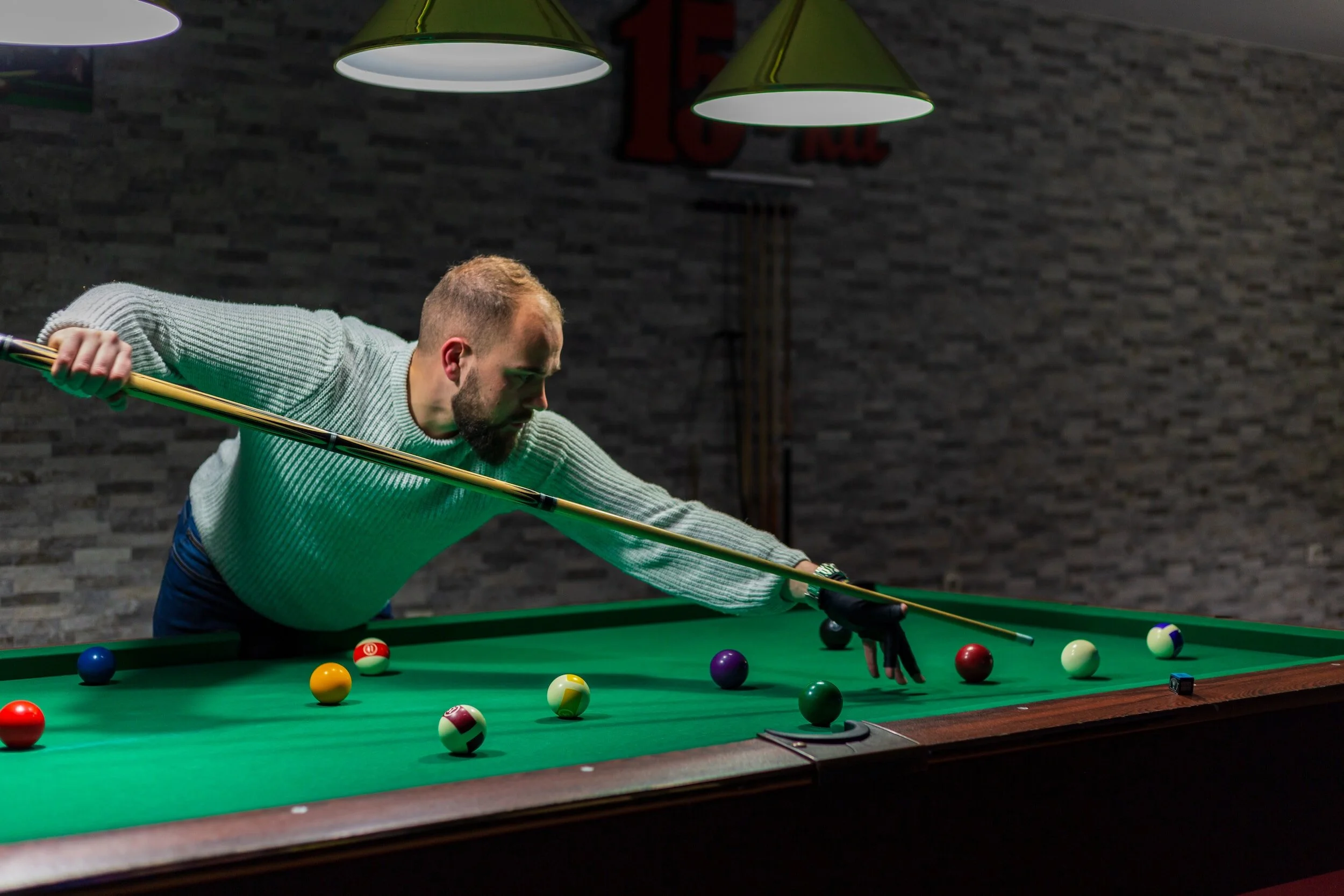 Man playing pool on a billiards table, lining up a shot with a cue stick, surrounded by colorful balls, in a dimly lit room with a brick wall background.