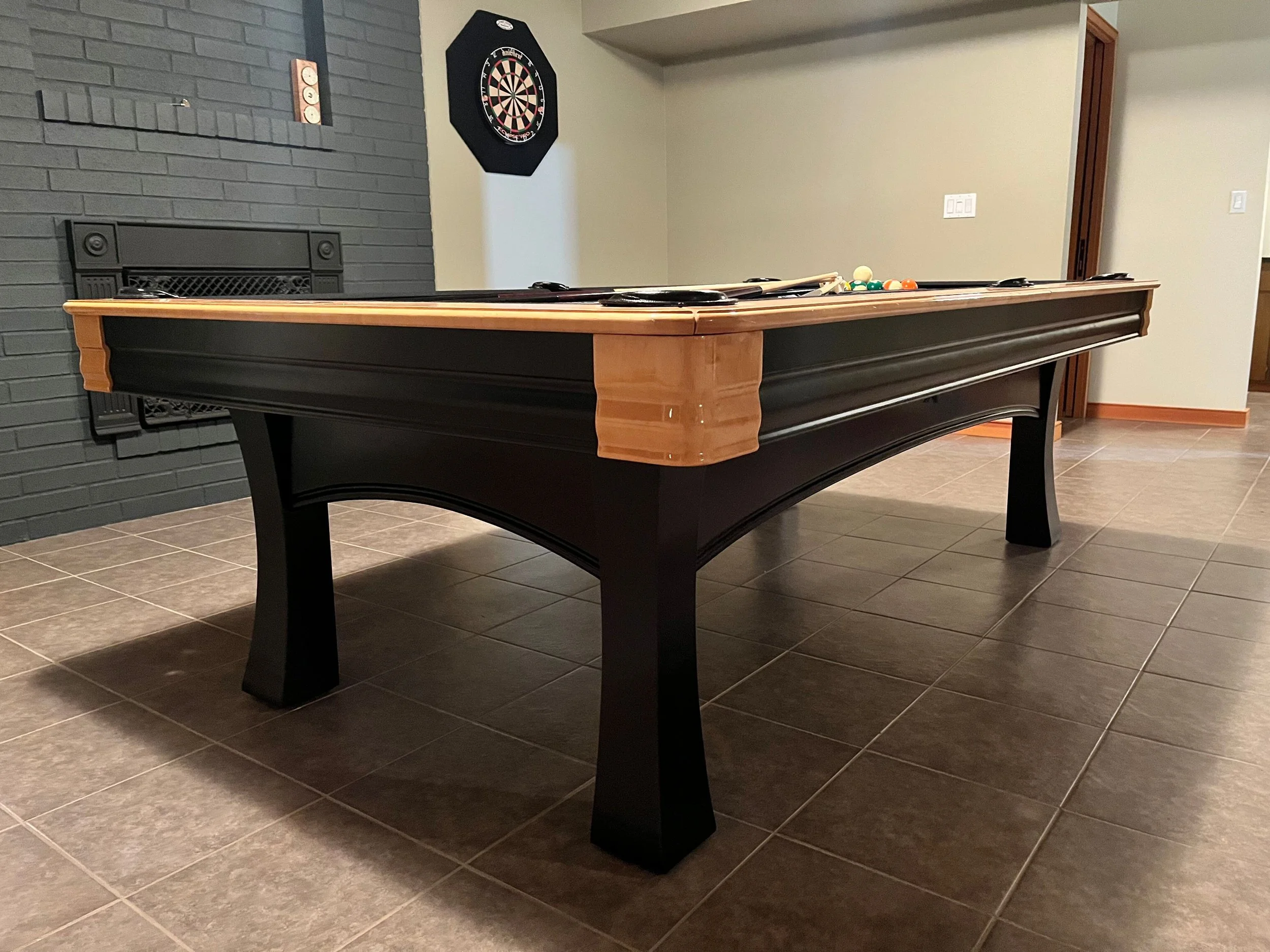 A black and wooden pool table in a basement with a wall-mounted dartboard and a fireplace.
