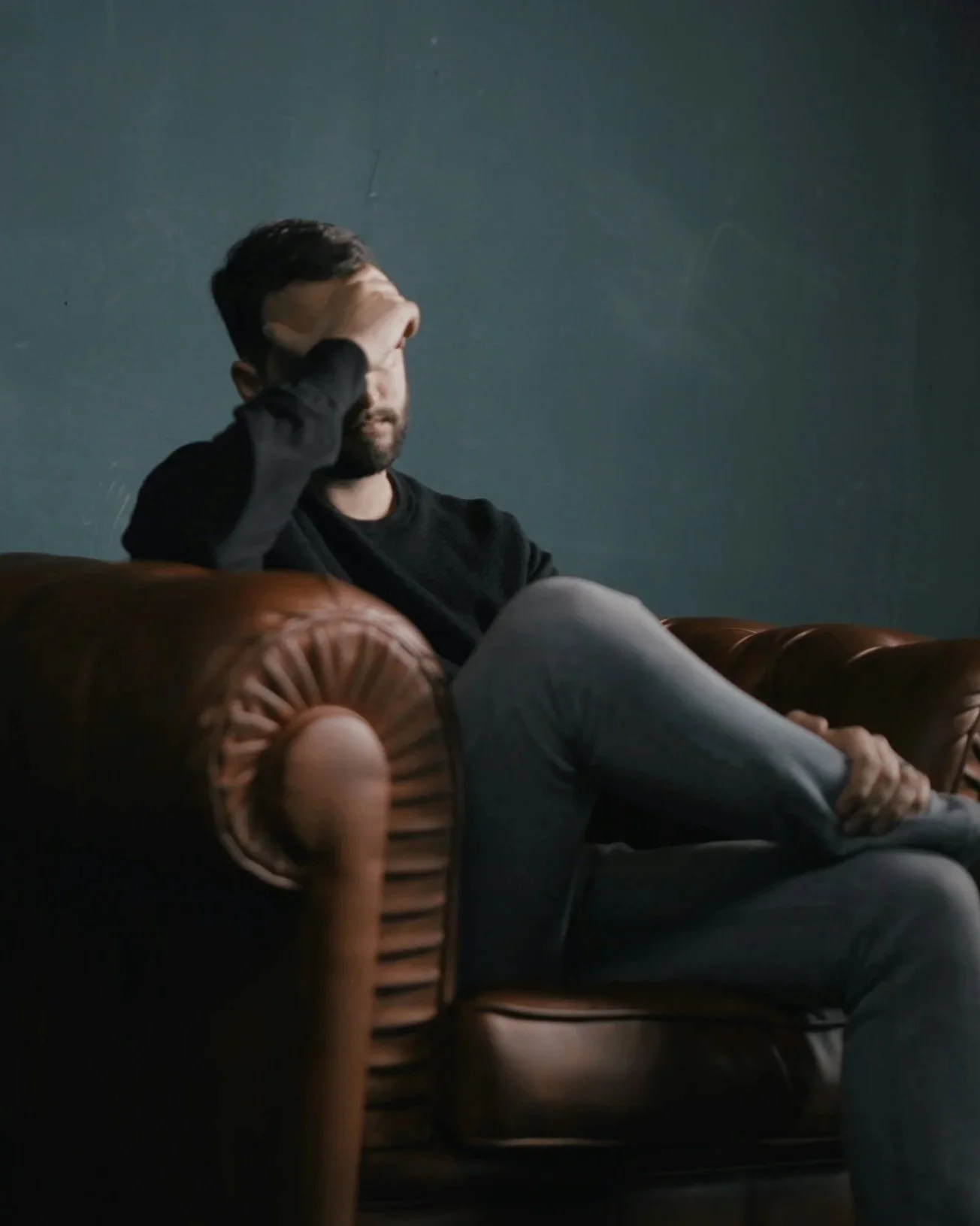 A man sitting on a vintage brown leather couch with one hand resting on his forehead and his eyes closed, in a room with a dark gray wall.