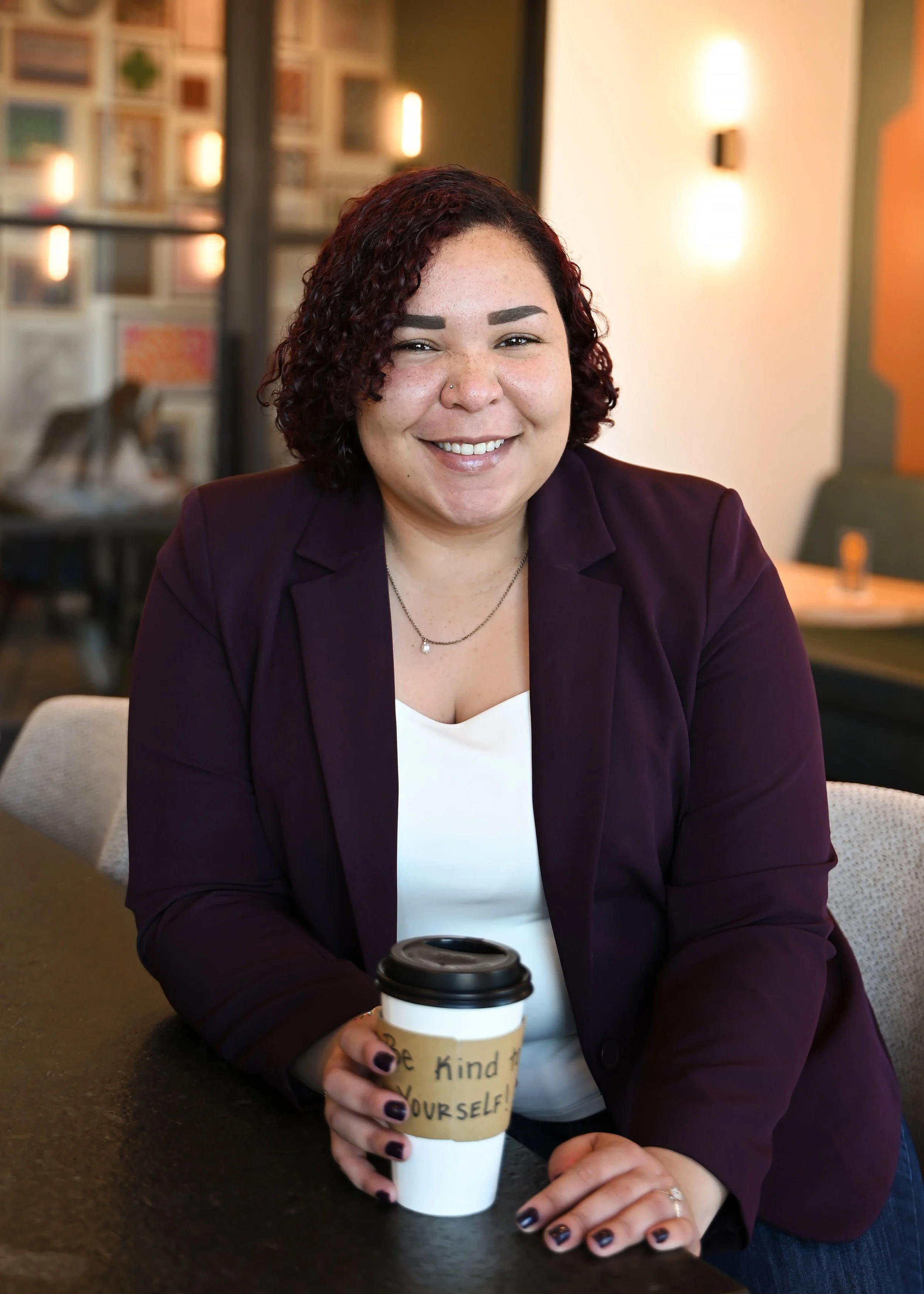 A woman with dark curly hair, wearing a dark purple blazer and a white top, smiling and holding a takeout coffee cup, sitting in a cozy cafe with warm lighting and decorative wall art. Kayla Storey-Silva, LPC Associate at Journey's Therapy Center.
