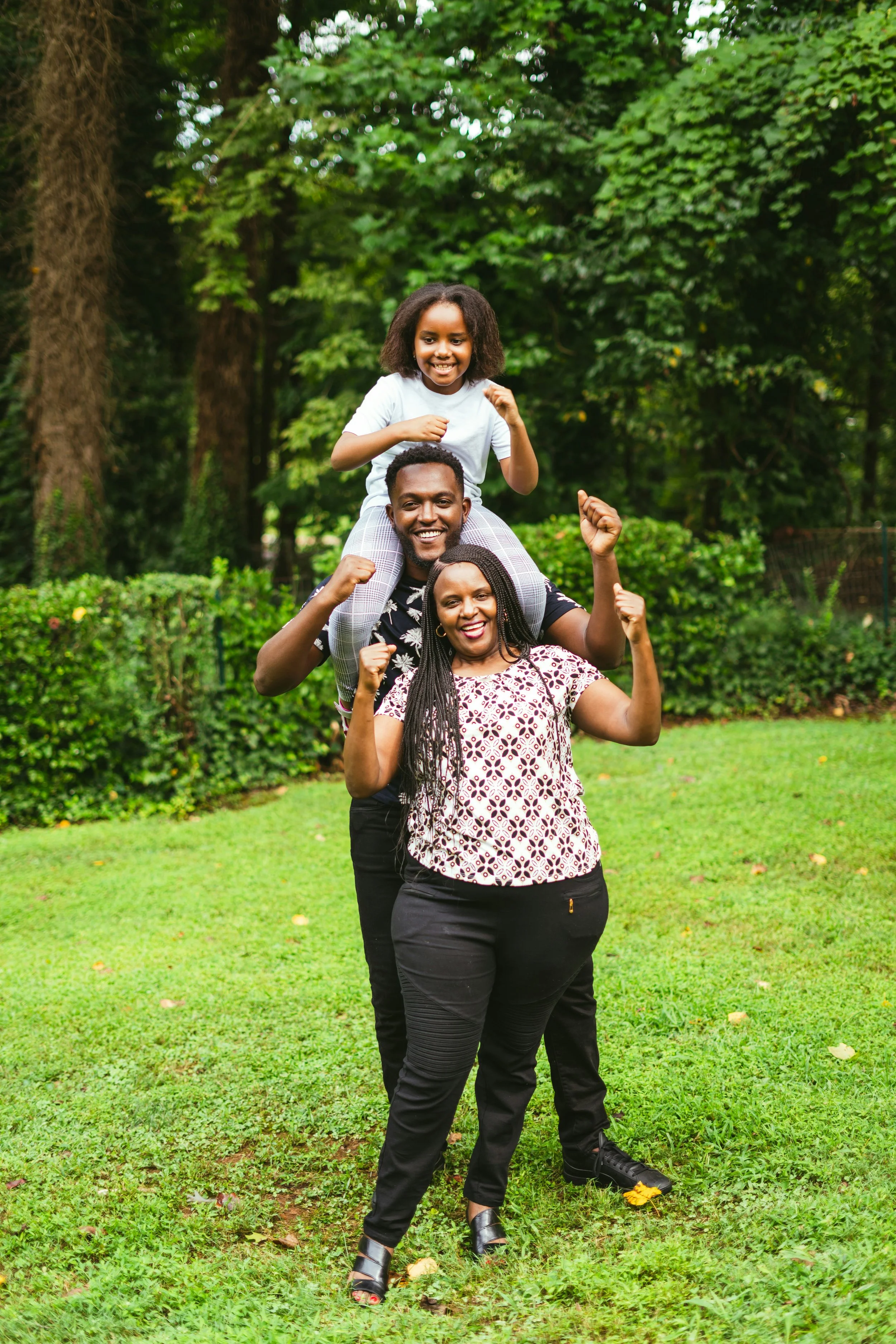 A family of three, a woman, a man, and a young girl, standing outdoors in a park with lush greenery. They are smiling and showing strength with their fists raised, with the man carrying the girl on his shoulders.
