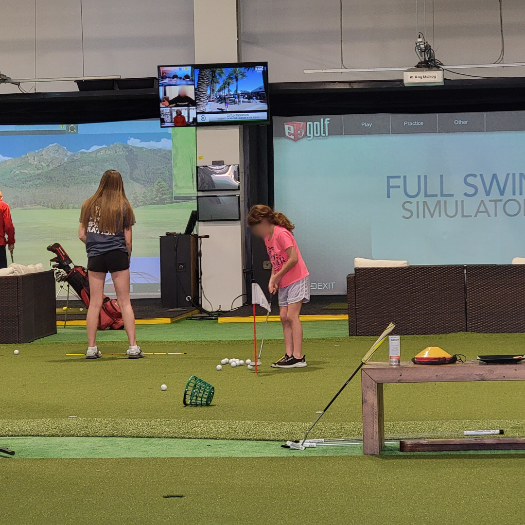 Indoor golf simulator with two girls practicing putting, one in a pink shirt and white shorts, and another in a gray shirt and black shorts, surrounded by golf balls and equipment.