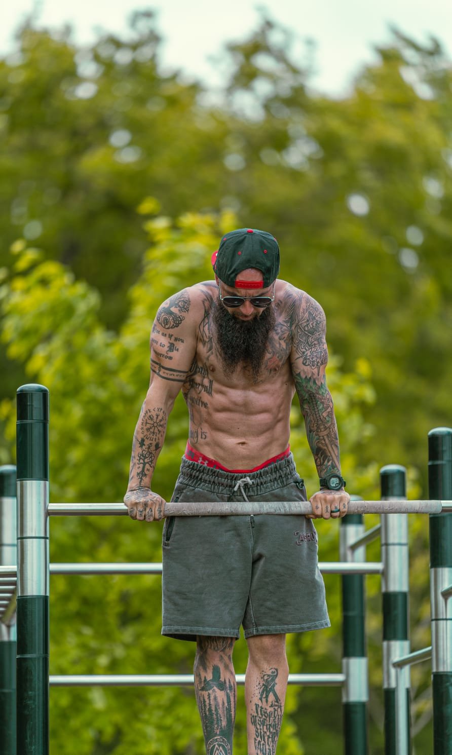A shirtless man with tattoos, wearing a black cap backwards, sunglasses, and shorts, doing a pull-up at an outdoor fitness park with green trees in the background.