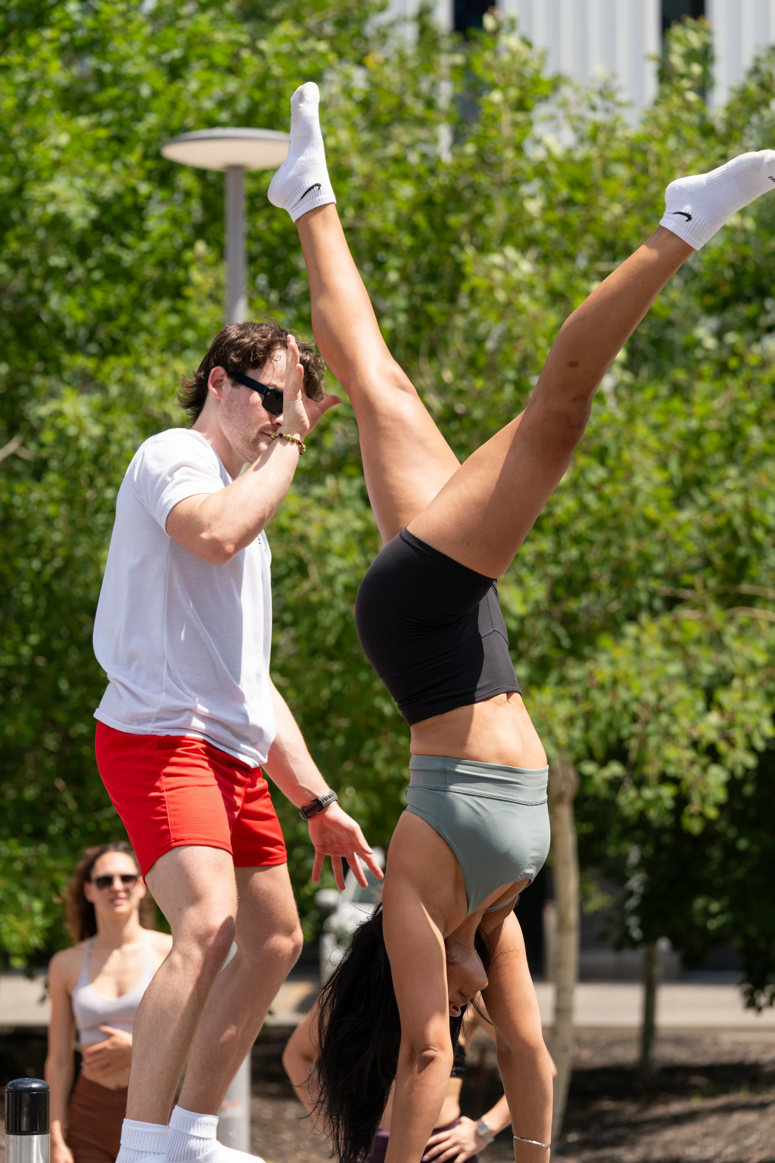 A person doing an acrobatic handstand outdoors on a sunny day, with spectators in the background. The performer is upside down, with legs extended upwards, wearing gray shorts and white socks. Another person, wearing sunglasses, a white T-shirt, and red shorts, is nearby.