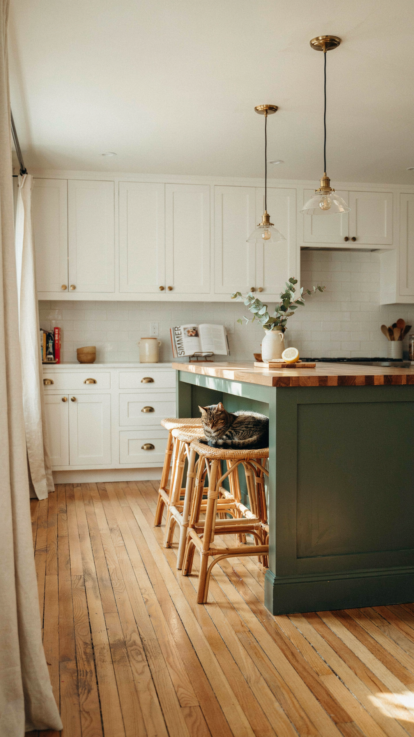 A bright cottage kitchen with white perimeter cabinets and a deep olive green painted kitchen island, a butcher block countertop on the island, honey-toned rattan bar stools, brass pendant lights overhead, wood plank floors, natural light,