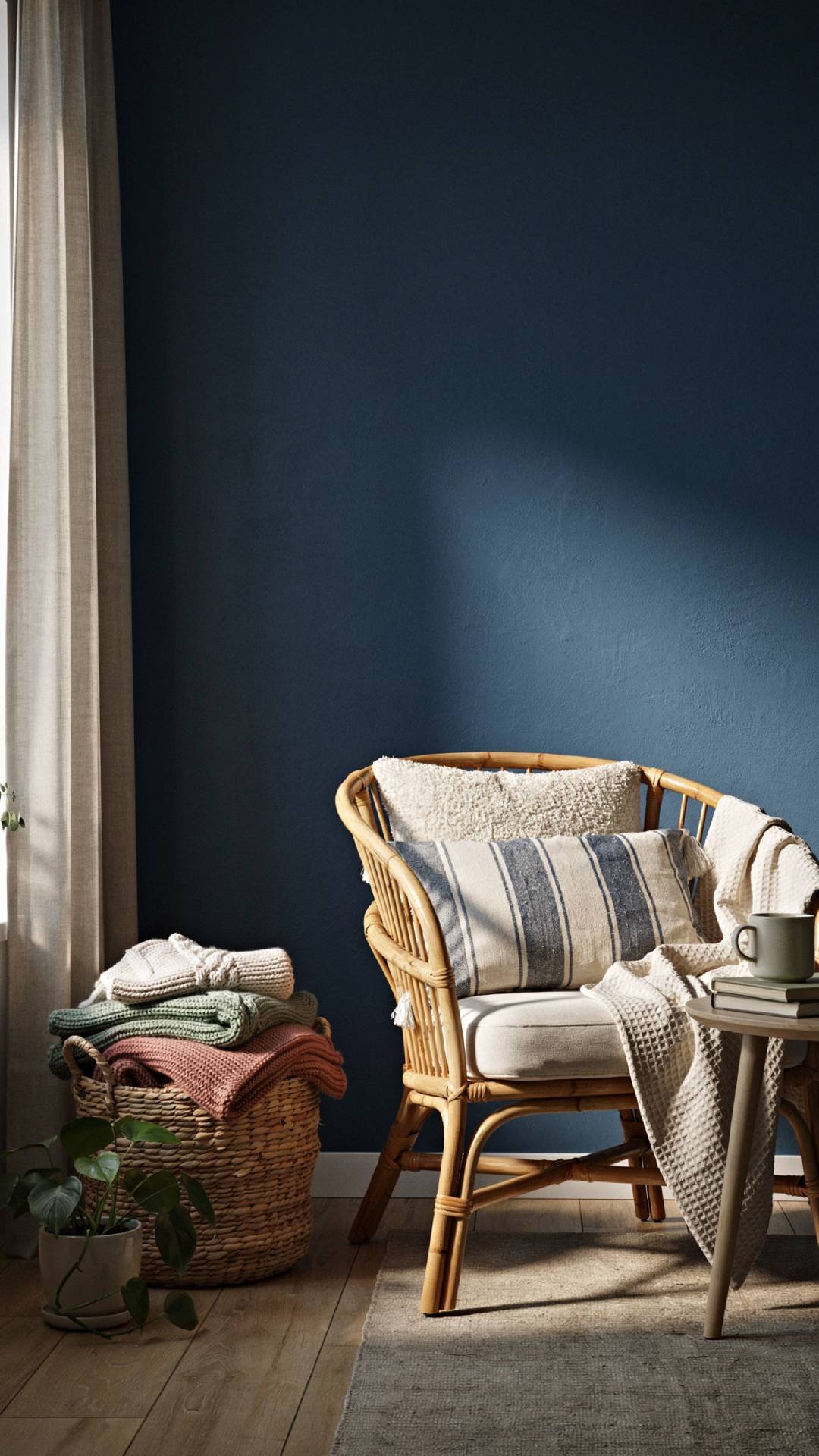 a cozy corner in a coastal living room with navy accent wall, rattan armchair layered with boucle and woven throw pillows, chunky waffle knit throw draped over the arm, seagrass basket of blankets on the floor, warm late-day light