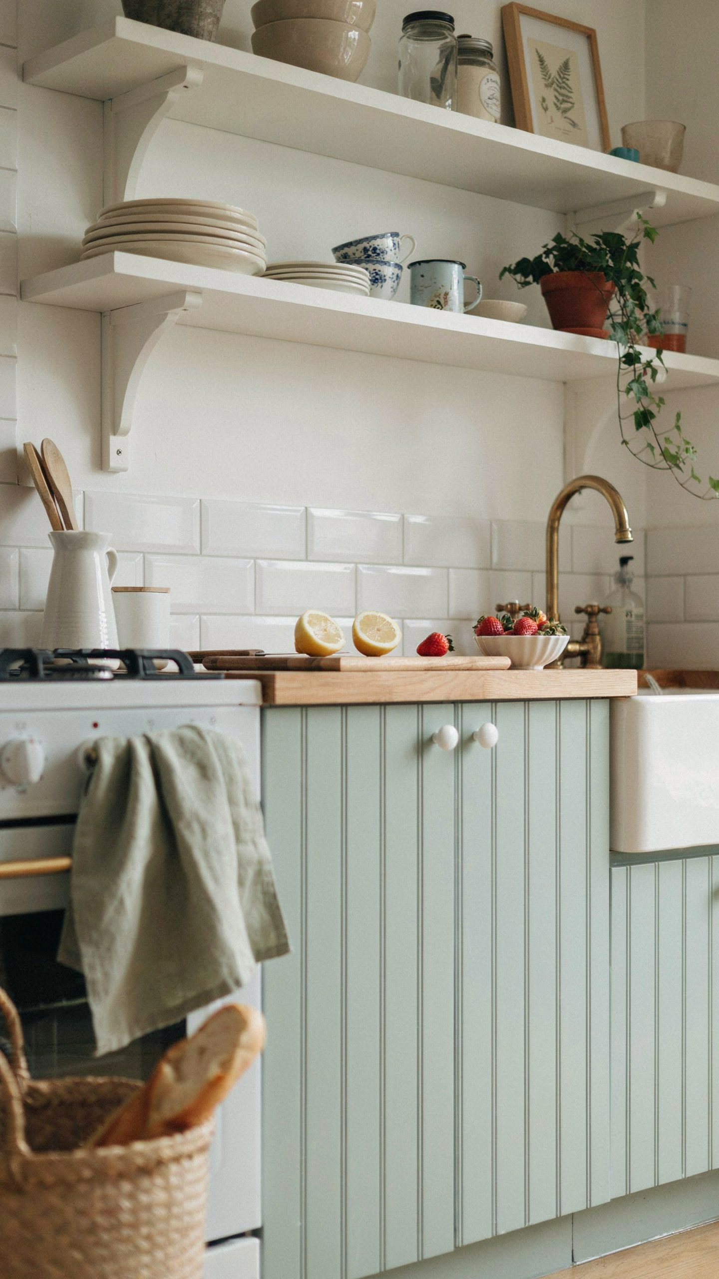 A vintage cottage kitchen with soft mint green beadboard cabinet doors, white ceramic knobs, white open floating wooden shelves above, a white subway tile backsplash, warm natural light, simple and charming farmhouse interior