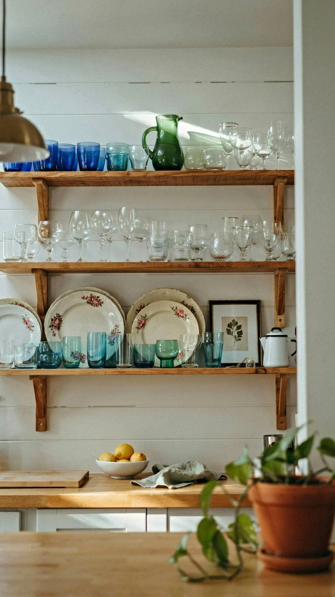 Full kitchen shot showing open wood shelving filled with mismatched blue and green glassware and vintage dishes, white shiplap wall behind the shelves, natural light catching the glass, warm and personal styling, photorealistic