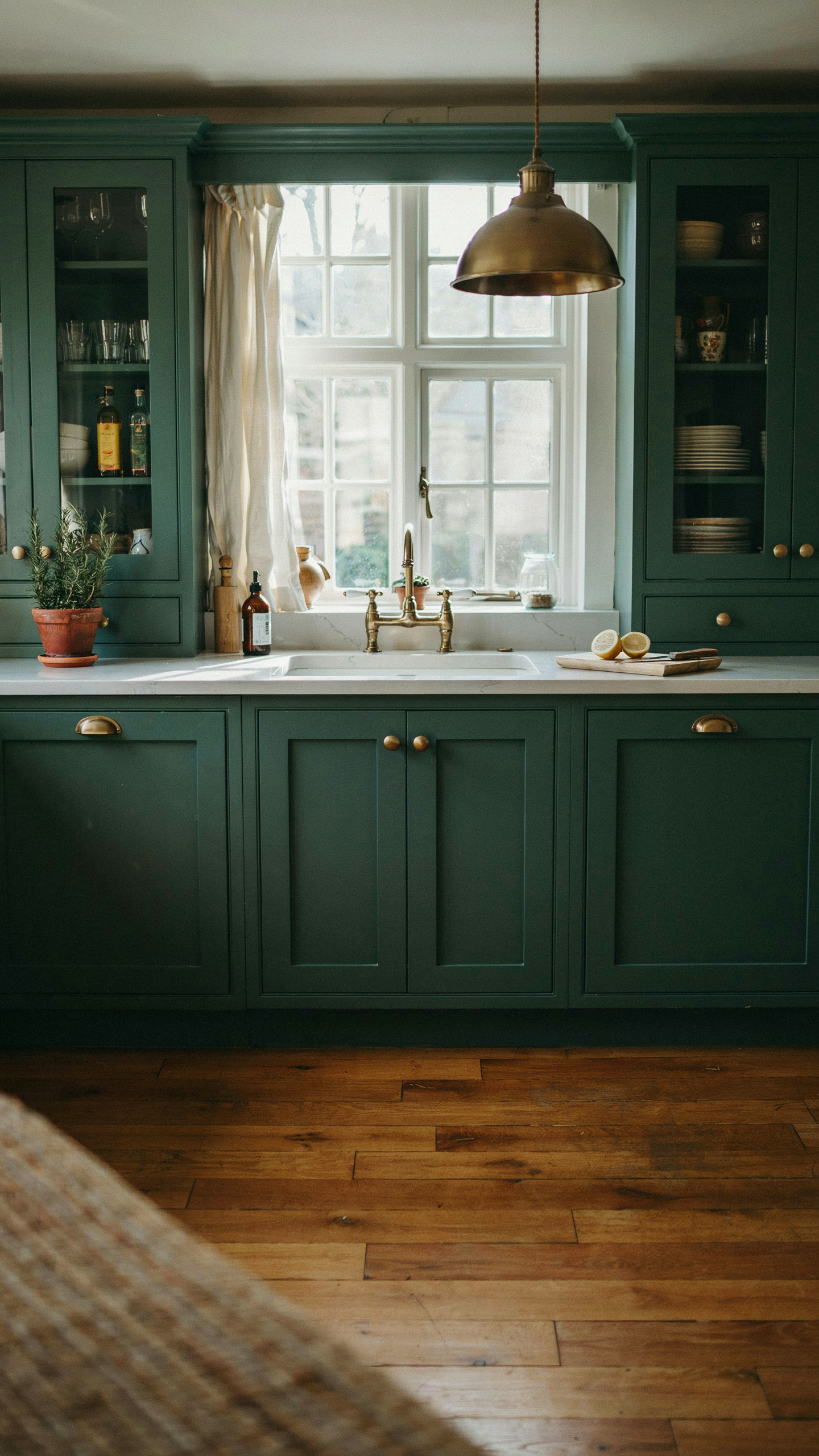 A moody and sophisticated cottage kitchen with deep forest green shaker cabinets, unlacquered brass hardware, a honed white quartz countertop, large windows letting in abundant natural light, glass-front upper cabinet doors, warm wood floors