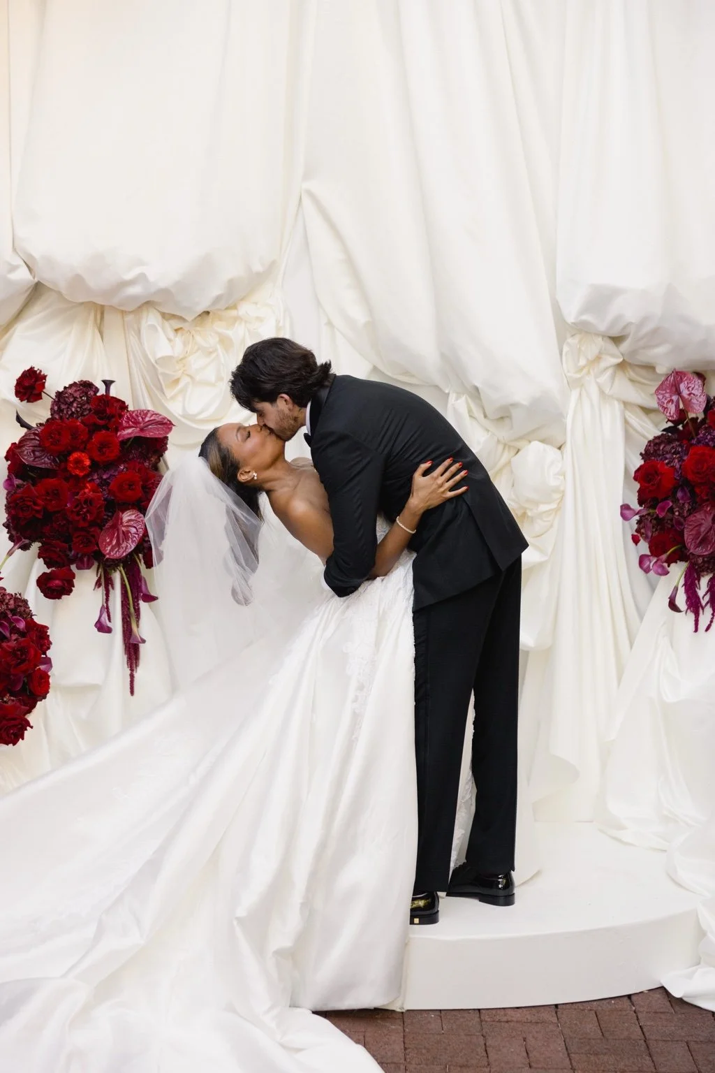 Taylor and Brandon share a wedding-day kiss surrounded by rich red florals and ivory drapery