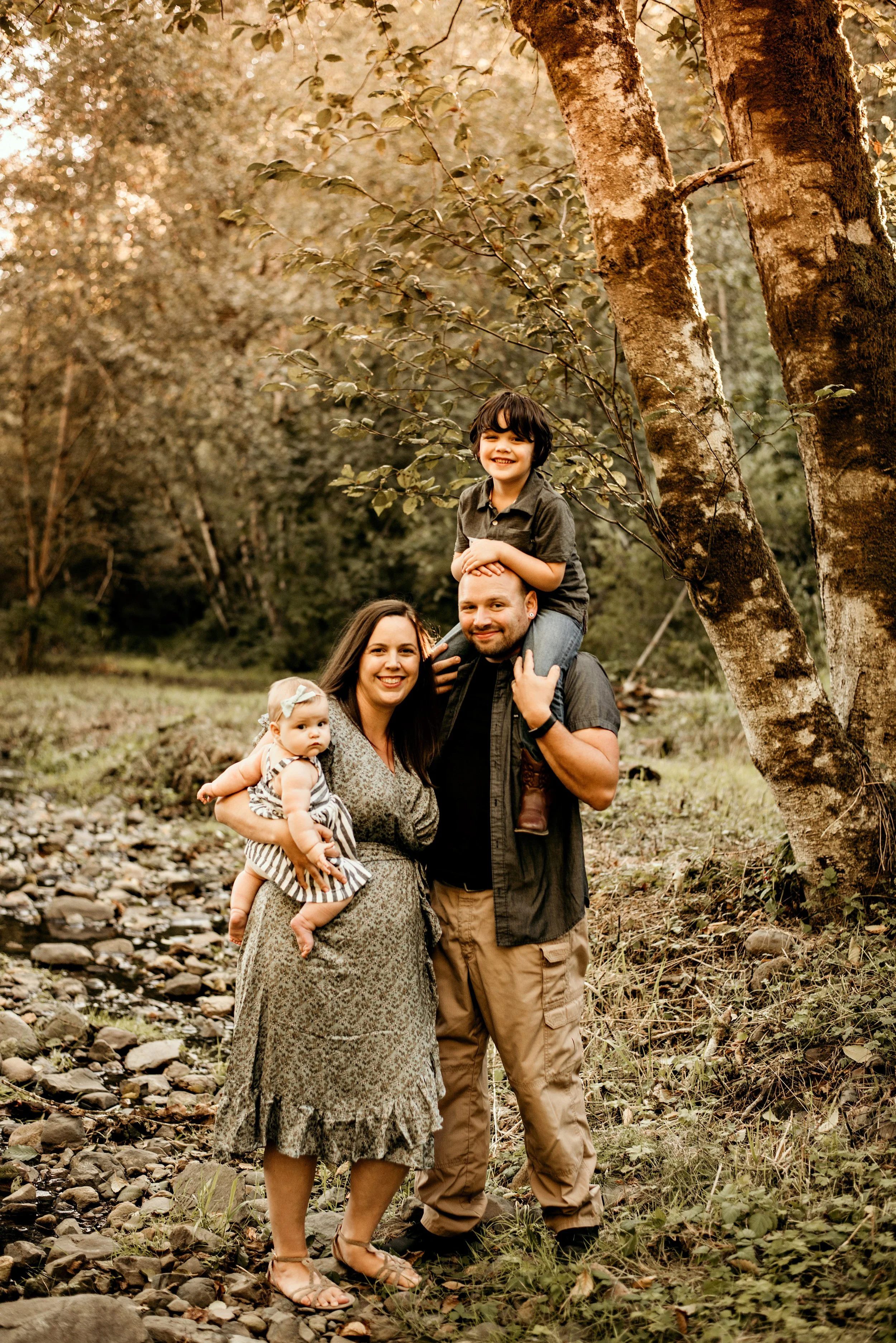 Dallas, OR, small business CPA and tax expert, Ashley, with family standing outdoors in a wooded area with trees and rocks, smiling at the camera. The mother holds a baby girl, and the father carries a young boy on his shoulders.