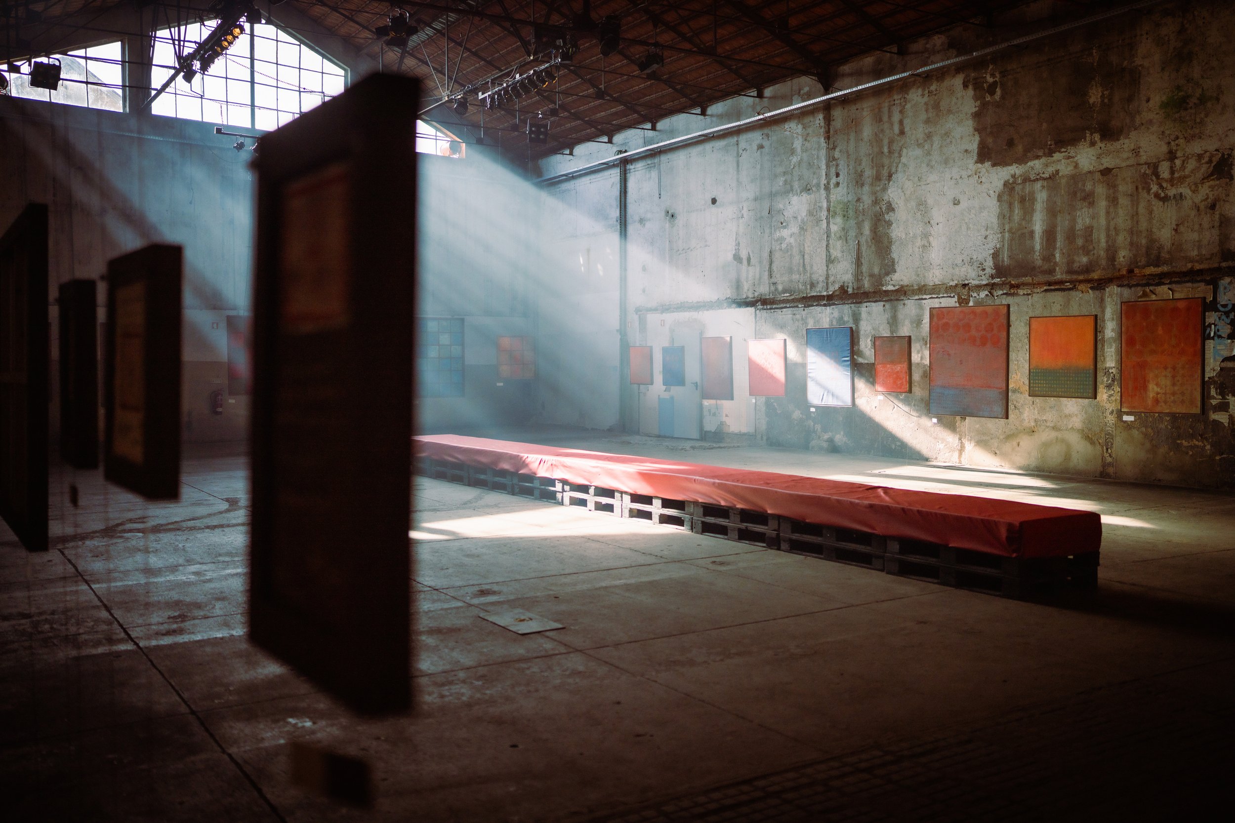 An indoor art gallery or exhibit space with paintings displayed on a concrete wall. Sunlight filters through large windows, creating beams of light inside the industrial-style room with a high, curved metal roof.