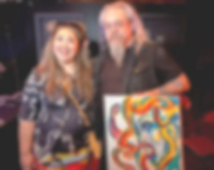 Two people smiling backstage at an event, with one holding a colorful abstract painting.