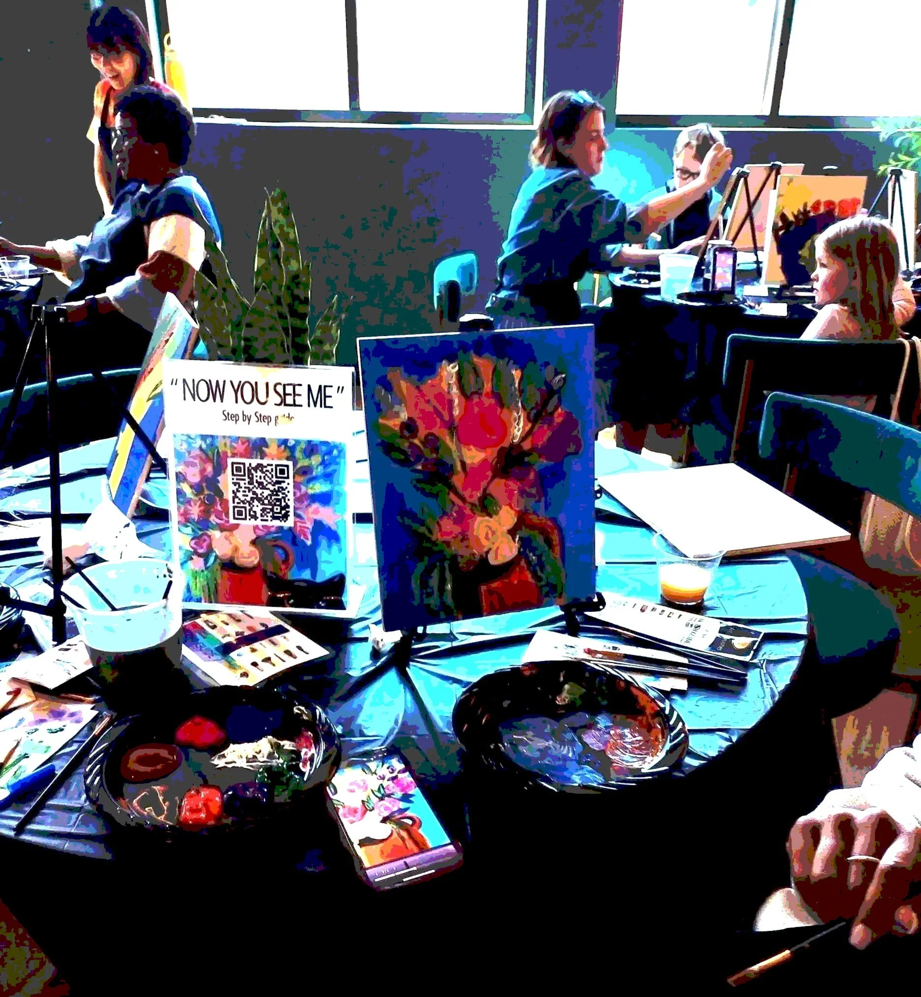 People sitting at tables in an art studio or classroom, painting on canvases, with large windows showing a cityscape and outdoor seating area.
