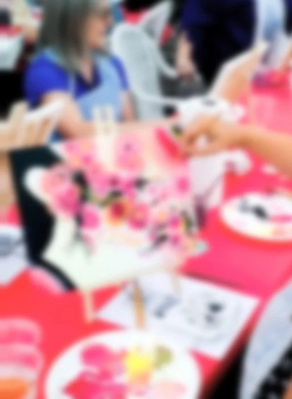 People participating in a painting or craft activity at a table filled with art supplies, with a focus on a colorful, decorated item in the foreground.