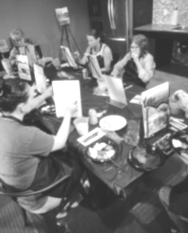 People participating in a painting or craft activity at a table filled with art supplies, with a focus on a colorful, decorated item in the foreground.