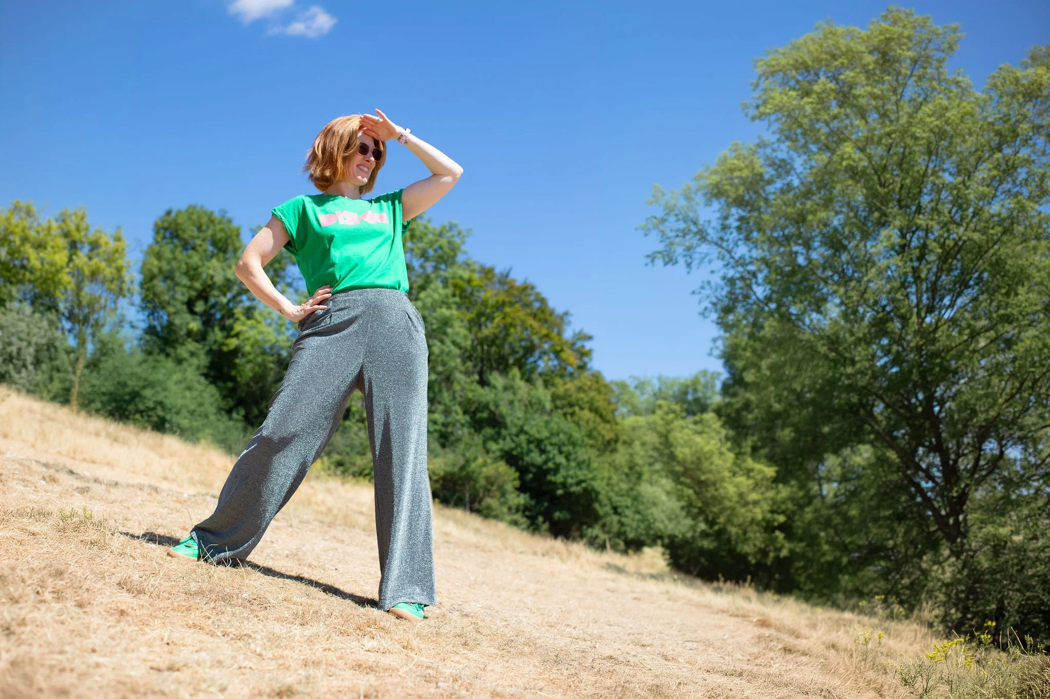 A woman with red hair, green t-shirt, silver glittery trousers, and teal shoes stands outdoors on a dry grassy hill, looking into the distance, shielding her eyes from the sun with her hand and smiling, with green trees and a bright blue sky behind.