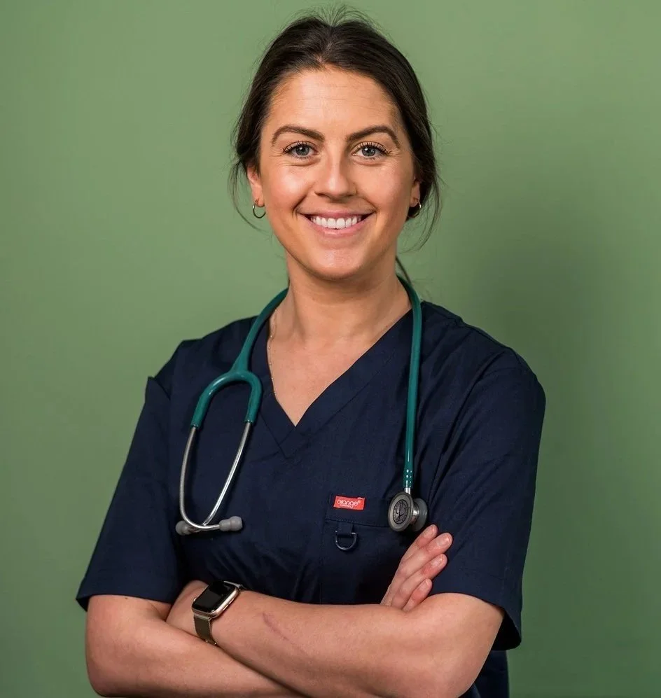 A smiling female healthcare professional wearing navy scrubs, a stethoscope around her neck, and a smartwatch on her wrist, standing against a green background.