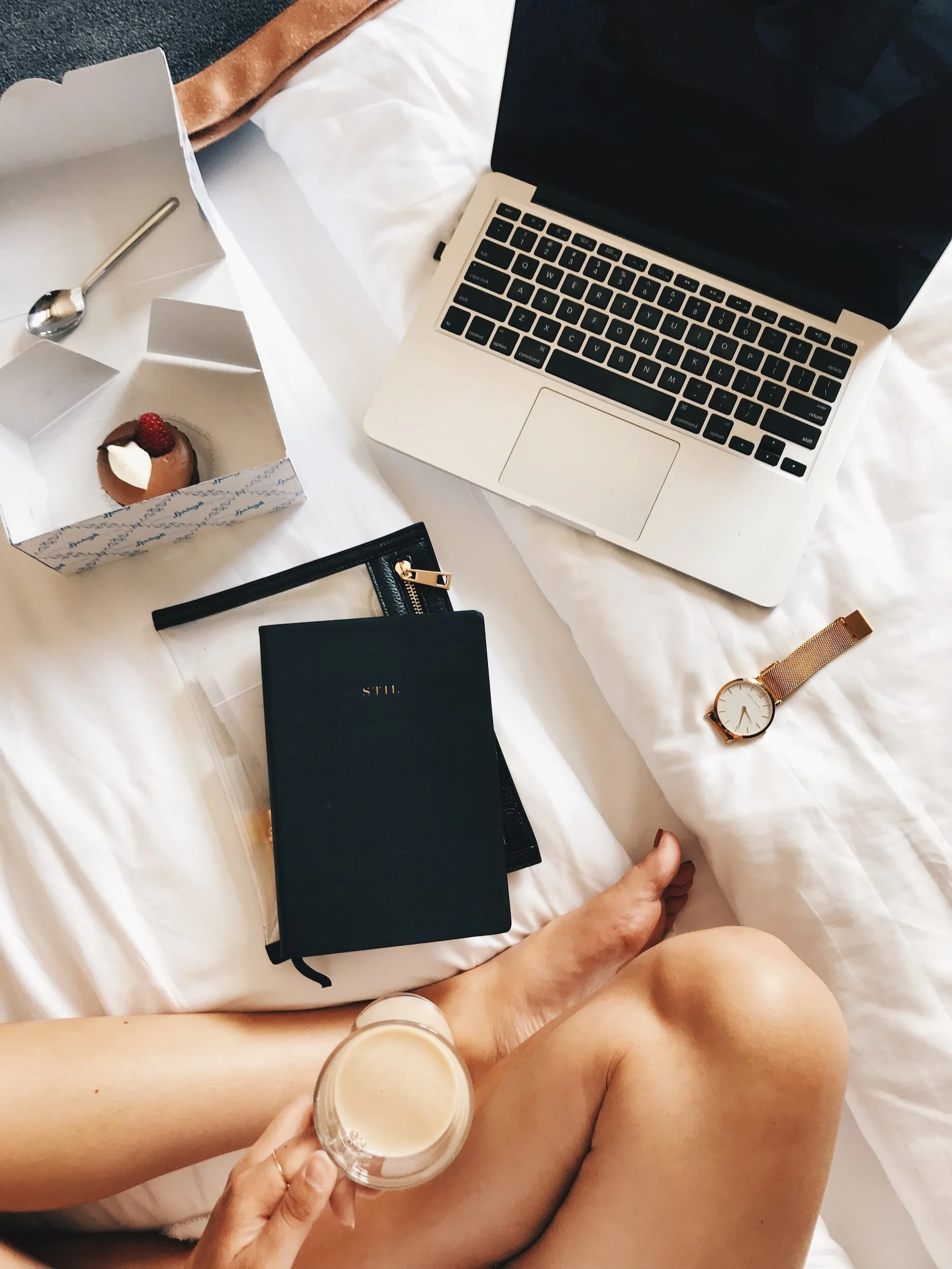 Image of a woman's legs on a bed with, books, a computer, a pastry, and the woman's hand holding a cup of coffee on top of her crossed legs.