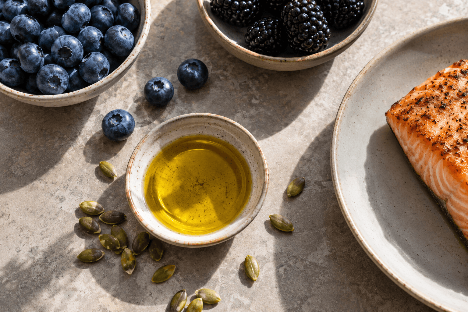 Overhead view of blueberries, blackberries, olive oil, pumpkin seeds, and salmon on a stone surface in natural light.