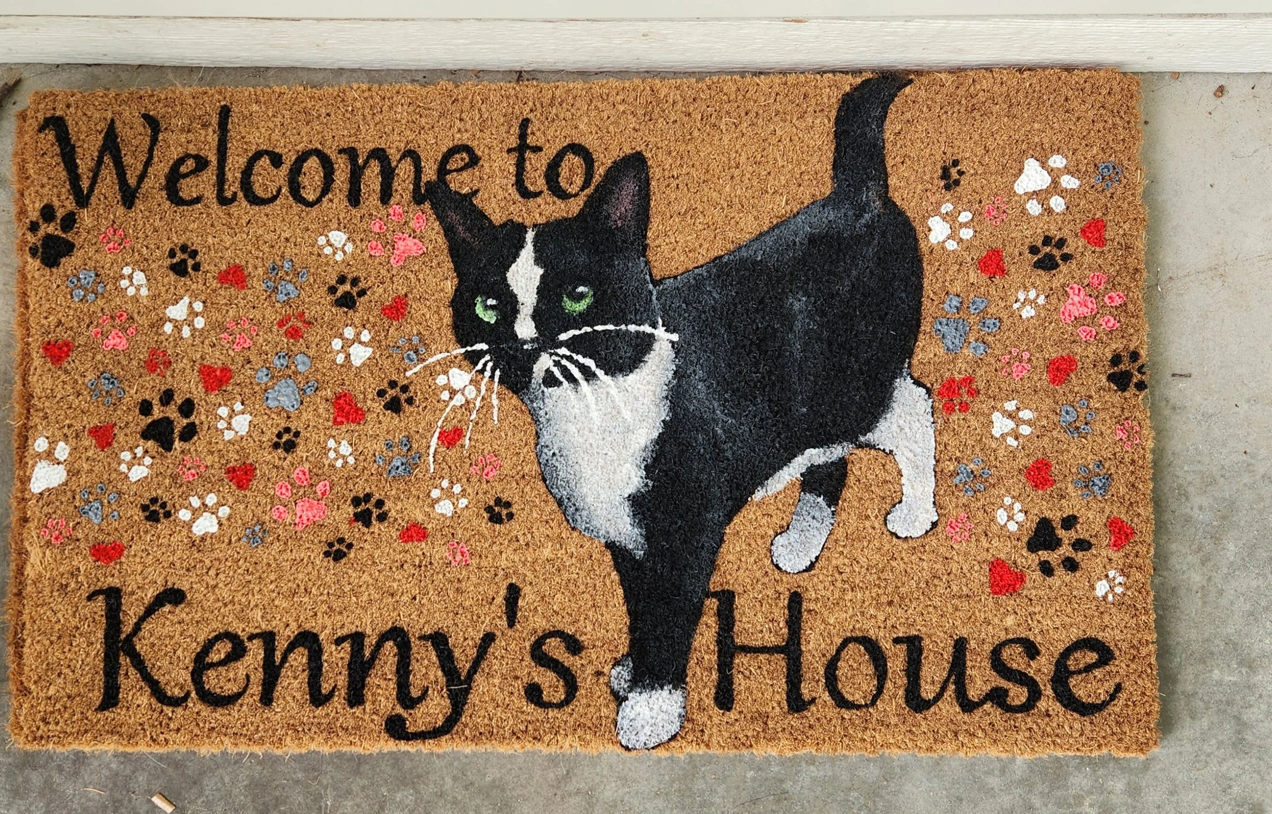 A welcome mat with a black and white cat, surrounded by colorful paw prints and hearts, and the text "Welcome to Kenny's House."