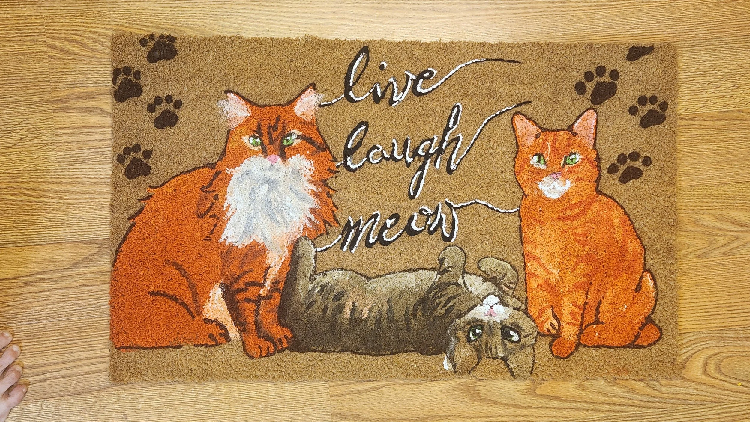 Decorative doormat featuring a large orange and white cat, an orange cat, and a gray tabby cat, with the words "live laugh meow" and paw print accents.