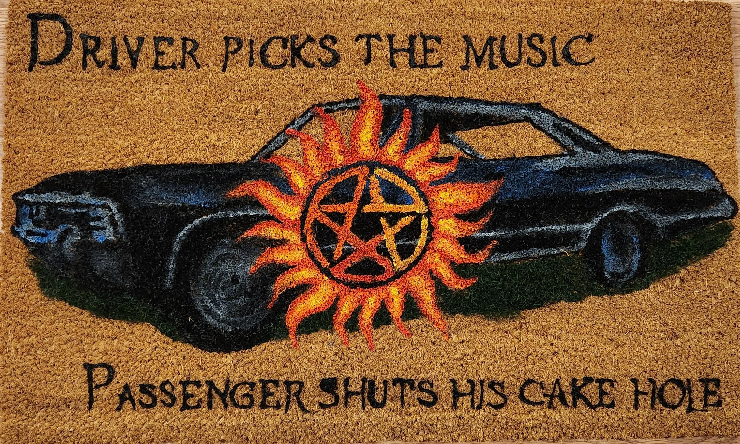A car decorated with a fiery sun and pentagram design, with the text "Driver picks the music, passenger shuts his cake hole".
