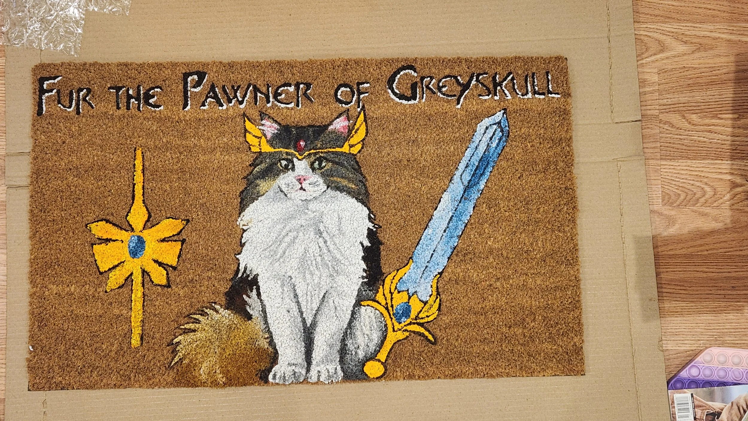 A decorative doormat with a cat wearing a crown, sword, and shield, and the text "Fur the Pawner of Greyskull."