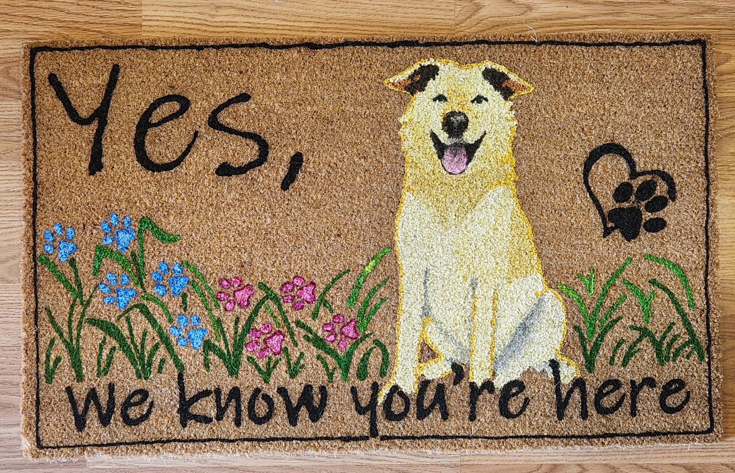 Doormat with a smiling dog, colorful flowers, a paw print, and the words "Yes, we know you're here"