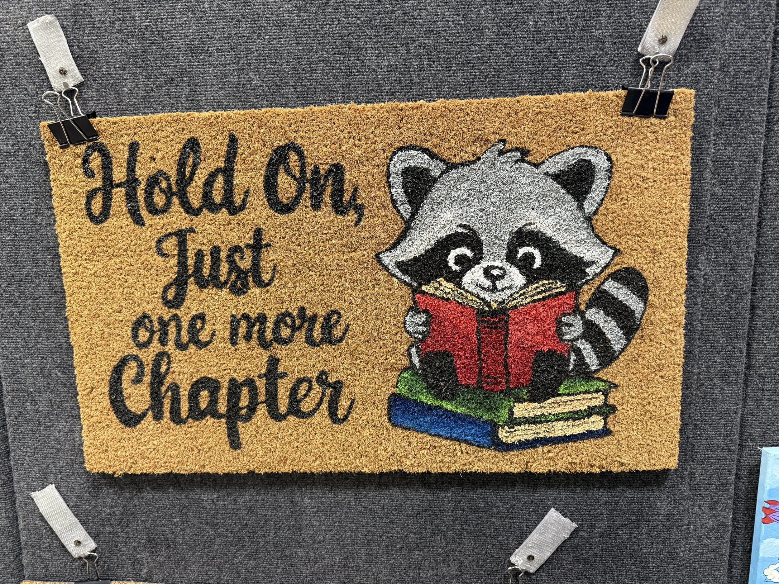 A decorative doormat featuring a raccoon reading a red book, sitting on three stacked books, with text that says, 'Hold On, Just one more Chapter'.