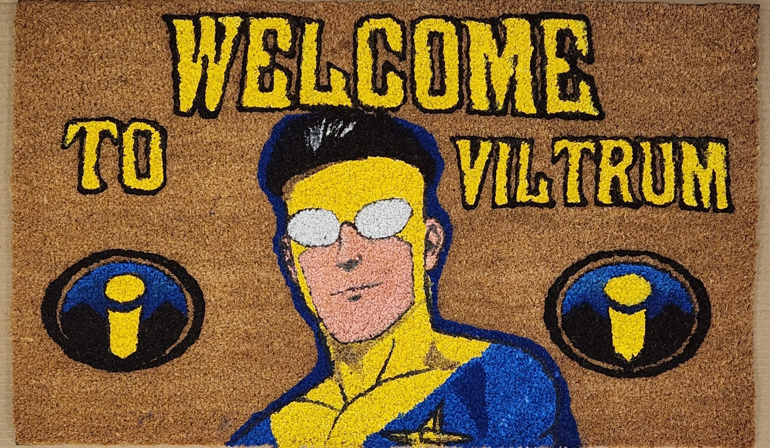 Welcome to Viltrum doormat with a cartoon illustration of a hero wearing glasses, a blue and yellow suit, and a confident expression, flanked by two icons resembling the Incredibles logo.