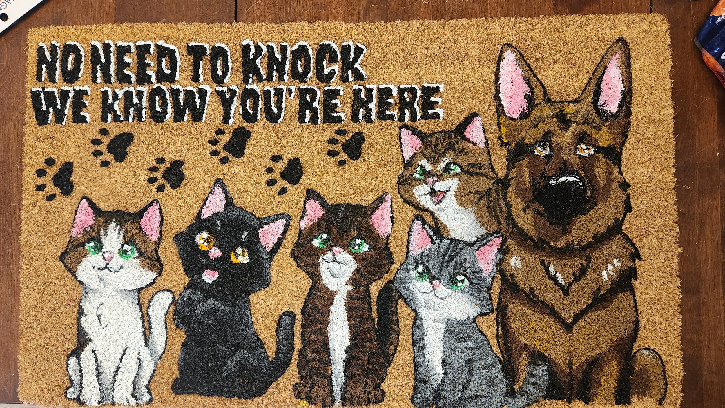 A doormat with a humorous design featuring four cats and a dog, with the text "No need to knock, we know you're here" above the animals.