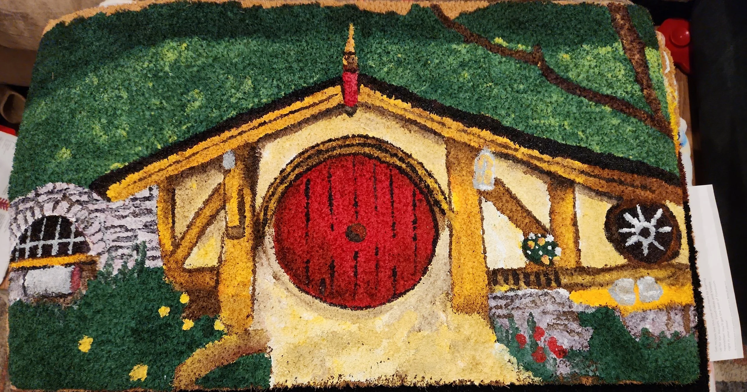 Colorful rug depicting a red barn with a round door, surrounded by green grass, flowers, and a stone wall.