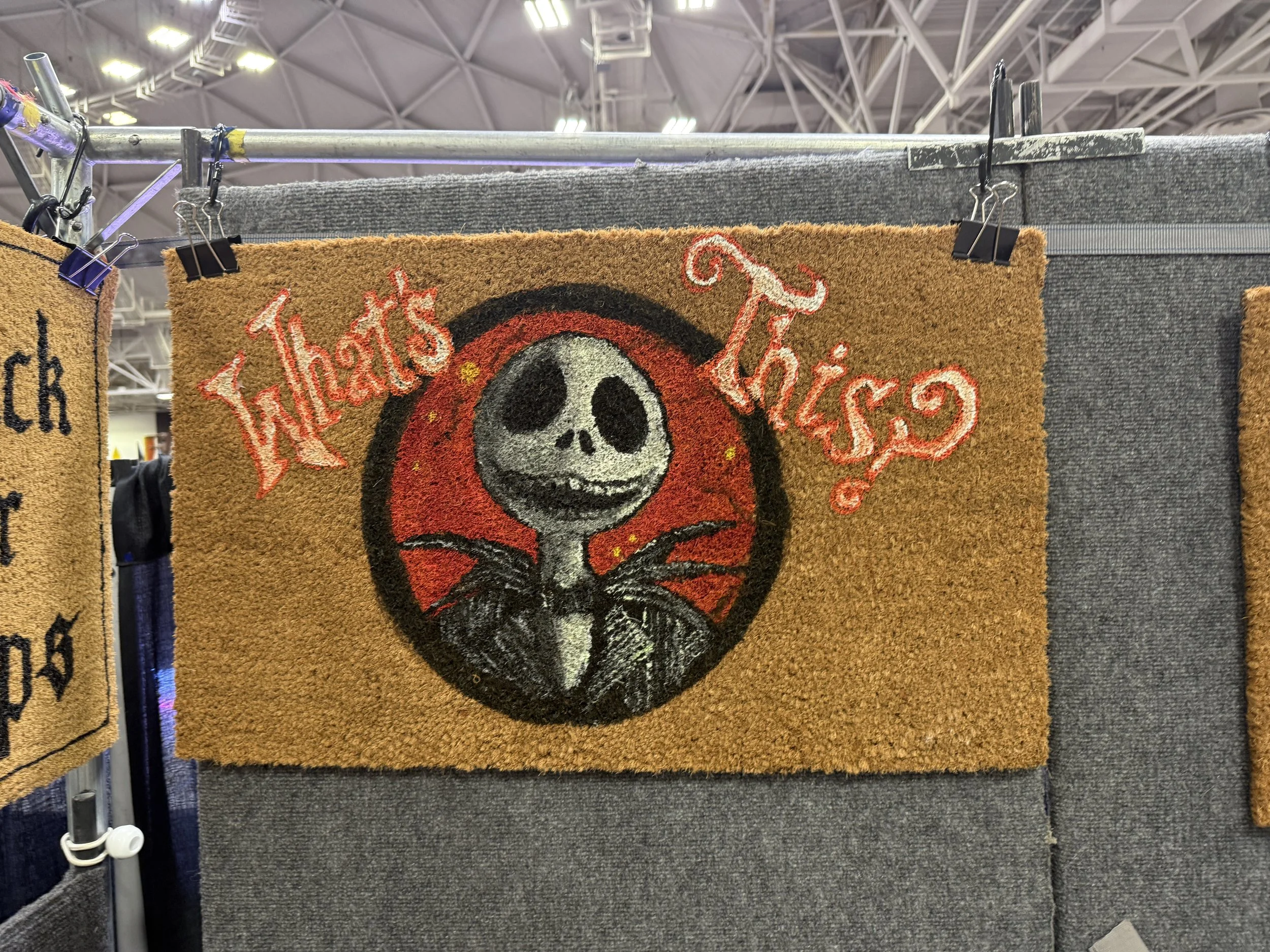 A decorative beige doormat features a stylized skeleton with a large skull and black eye sockets, set against a red moon and black wings. Above the skeleton, pink cursive text reads 'What's This?'