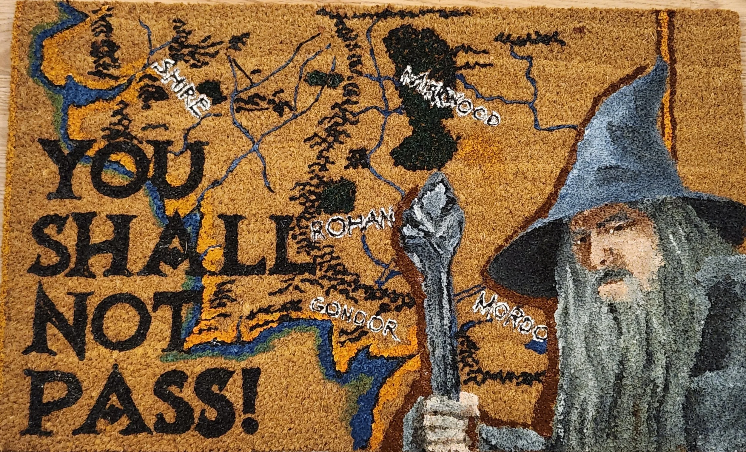 A woven tapestry or wall hanging with a map of Middle-earth from The Lord of the Rings, showing Rohan, Gondor, and Mordor. Text on the tapestry reads, "You shall not pass!" A depiction of Gandalf the Grey wearing a grey hat and robe, holding a staff.
