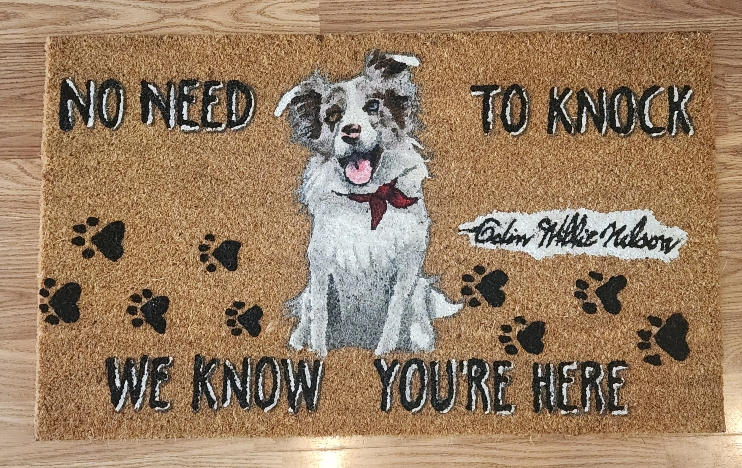 A welcome doormat featuring a cute cartoon dog with a red bandana. The mat has the text 'No need to knock, we know you're here' and includes paw print designs.