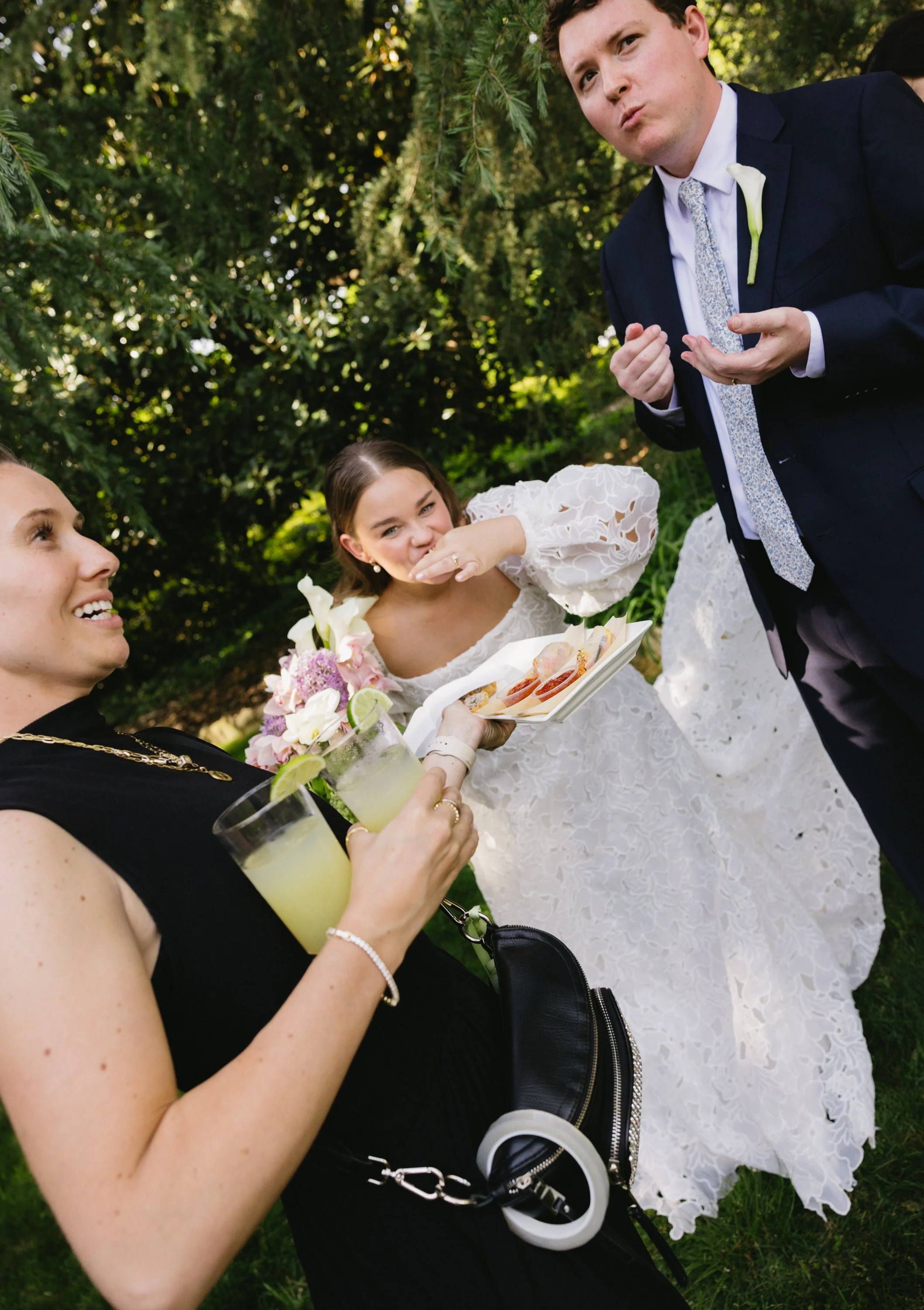 Two women and a man at an outdoor wedding reception. One woman in a black dress holds two drinks with lime wedges. The other woman in a white wedding dress holds a bouquet and eats at a small platter. The man, in a dark suit, is standing and gesturin