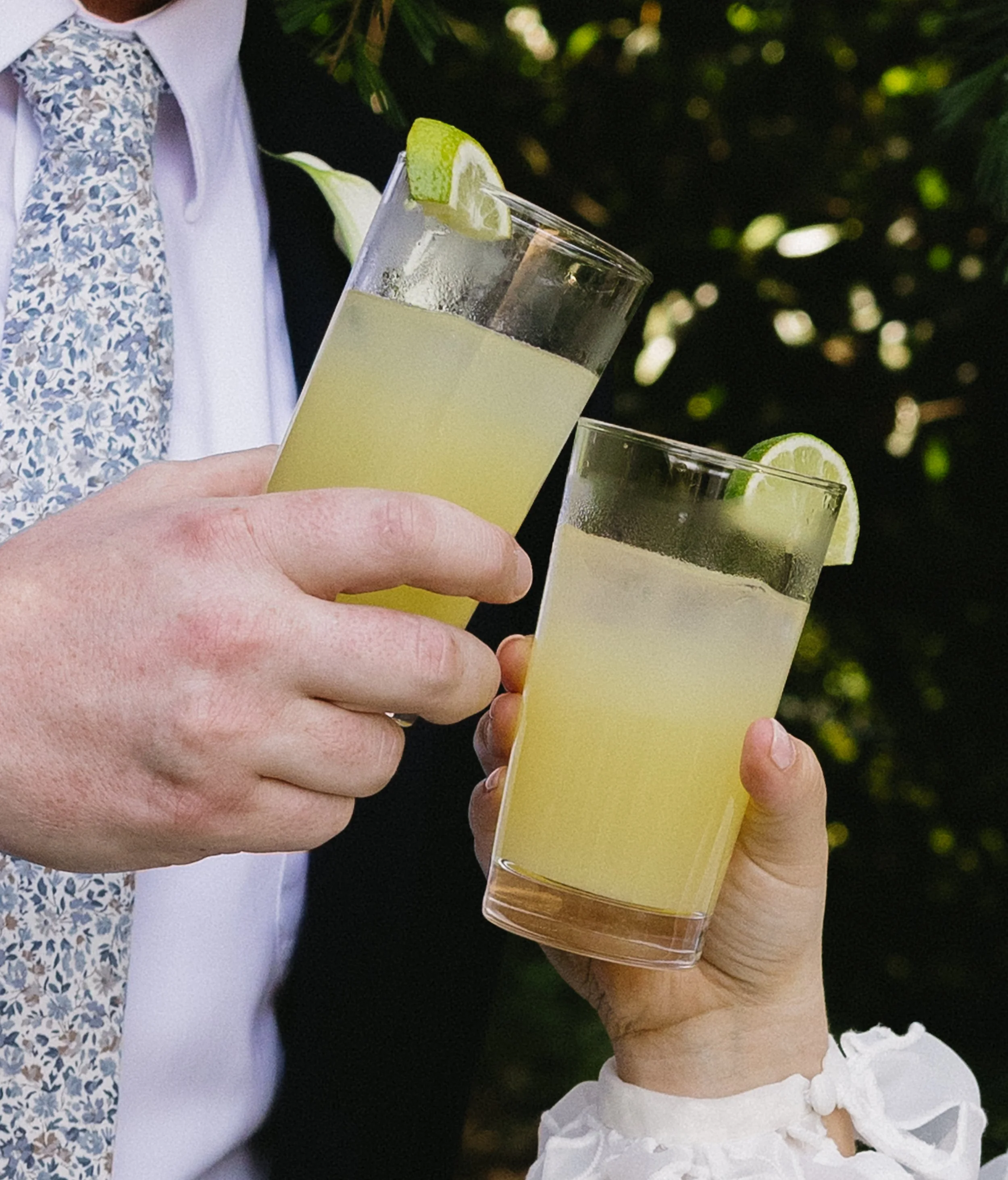 Two people holding glasses of lemonade with lime wedges, toasting outdoors in front of greenery.